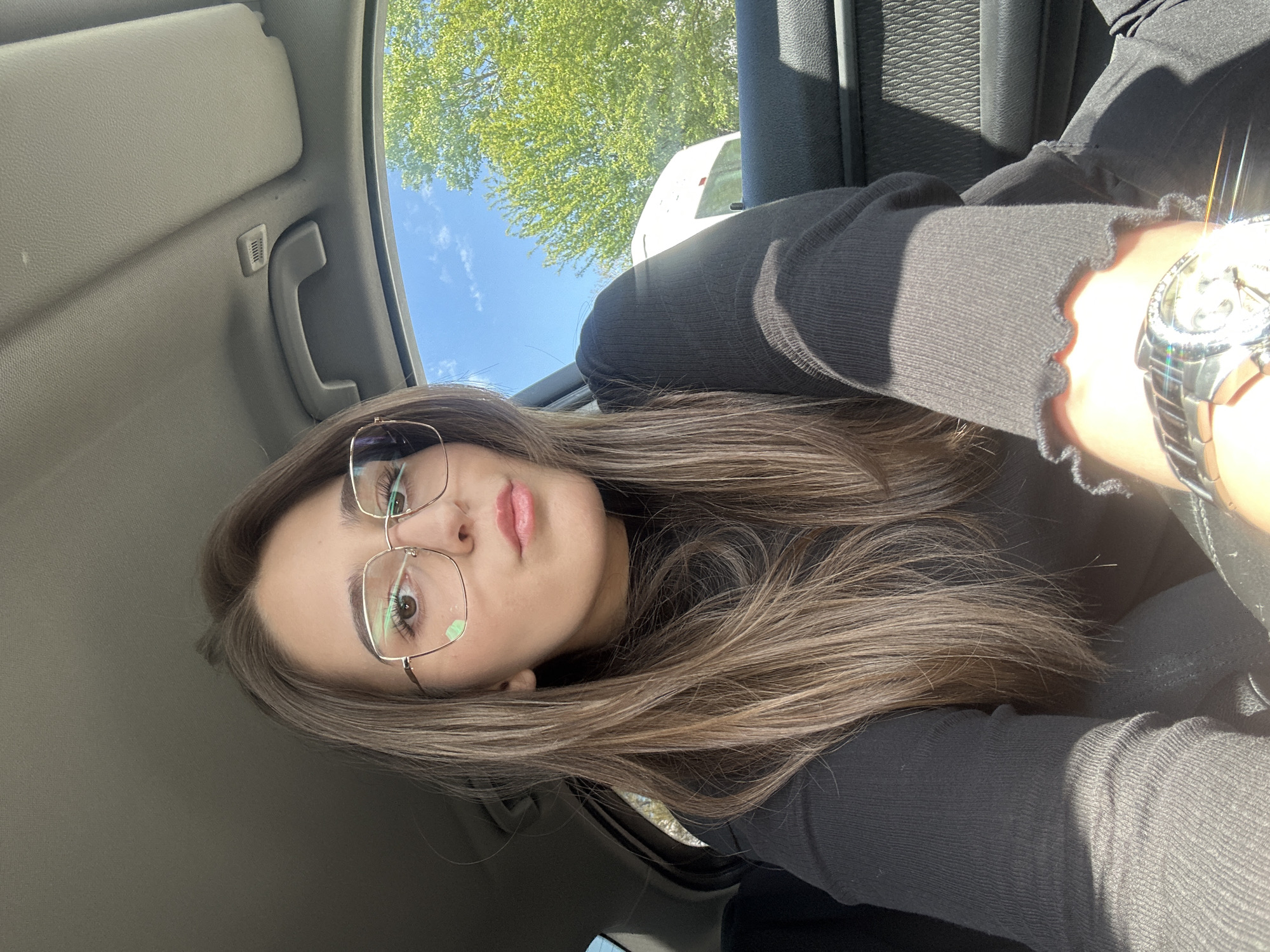 A woman with long brown hair and glasses sits inside a car. She wears a black shirt and a watch on her left wrist. Outside the car window, green trees and a patch of blue sky are visible. A white vehicle is partially visible. The location is likely in a car in Sofia, Bulgaria, although this is not explicitly indicated.