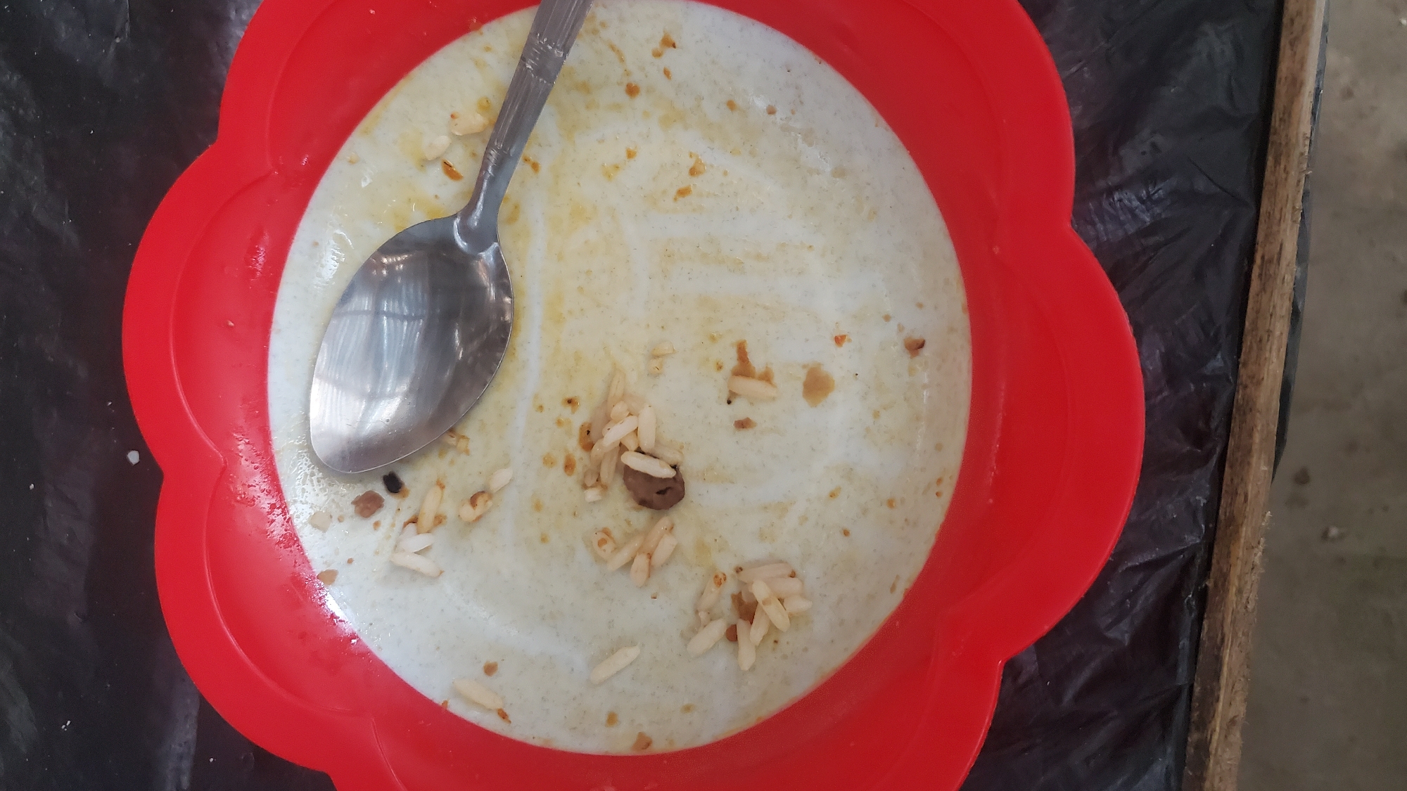 An overhead shot shows a used, dirty red plastic plate with a silver spoon resting on it. The plate contains remnants of rice and sauce. The plate is placed on a black plastic surface which is on a wooden surface. The scene appears to be outdoors in Jalingo, Nigeria, given the lighting and general environmental conditions.