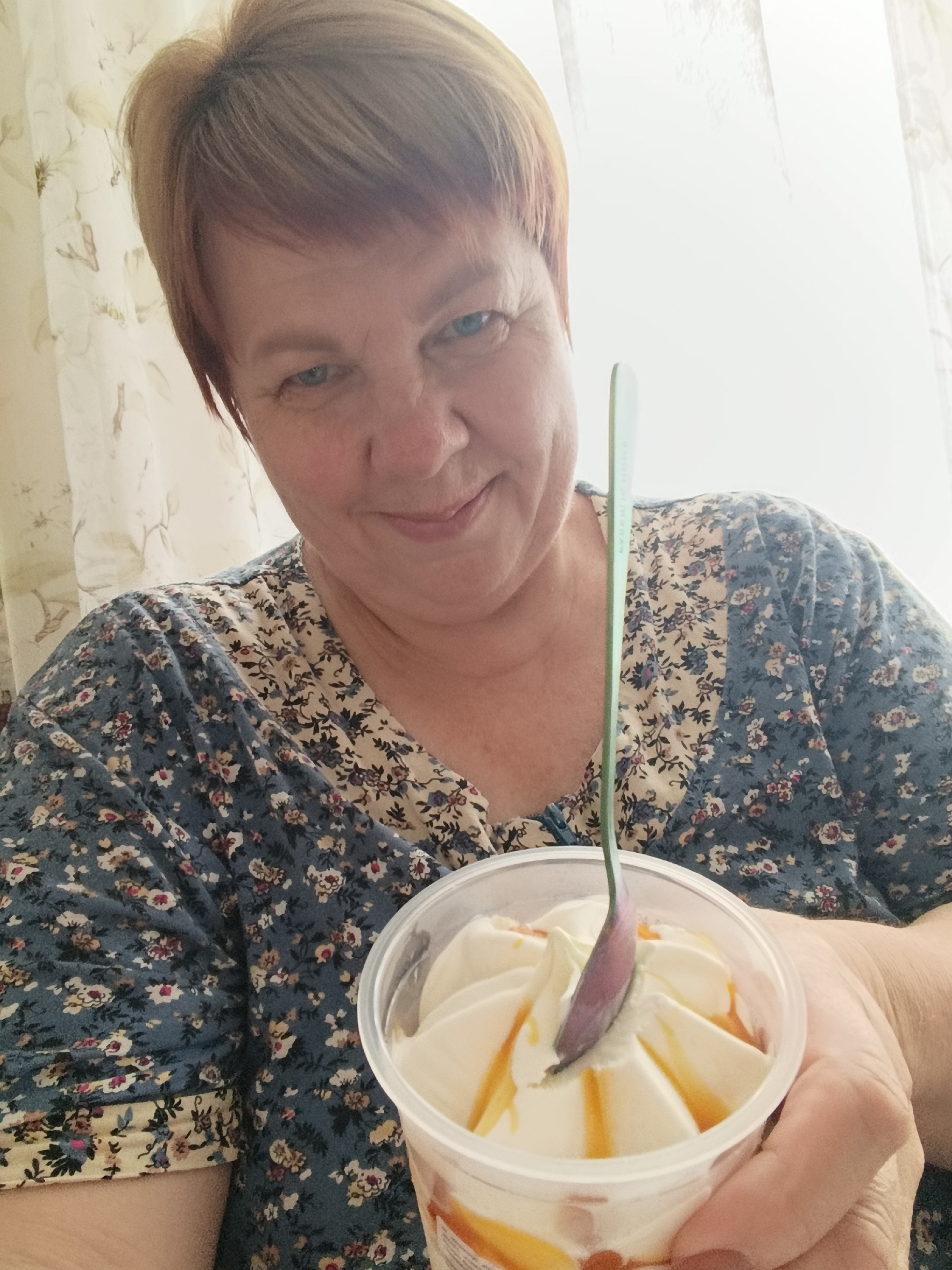 An eye-level, medium shot captures a woman with short, reddish-blonde hair and blue eyes, holding a clear plastic cup filled with soft-serve ice cream topped with caramel sauce. She is wearing a blue and cream floral-patterned shirt and appears to be smiling as she looks down at the ice cream. A light green spoon is partially submerged in the ice cream. The background consists of a light-colored curtain with a floral pattern. The scene is likely indoors, possibly in an apartment, in the city of Svietlahorsk, Belarus.