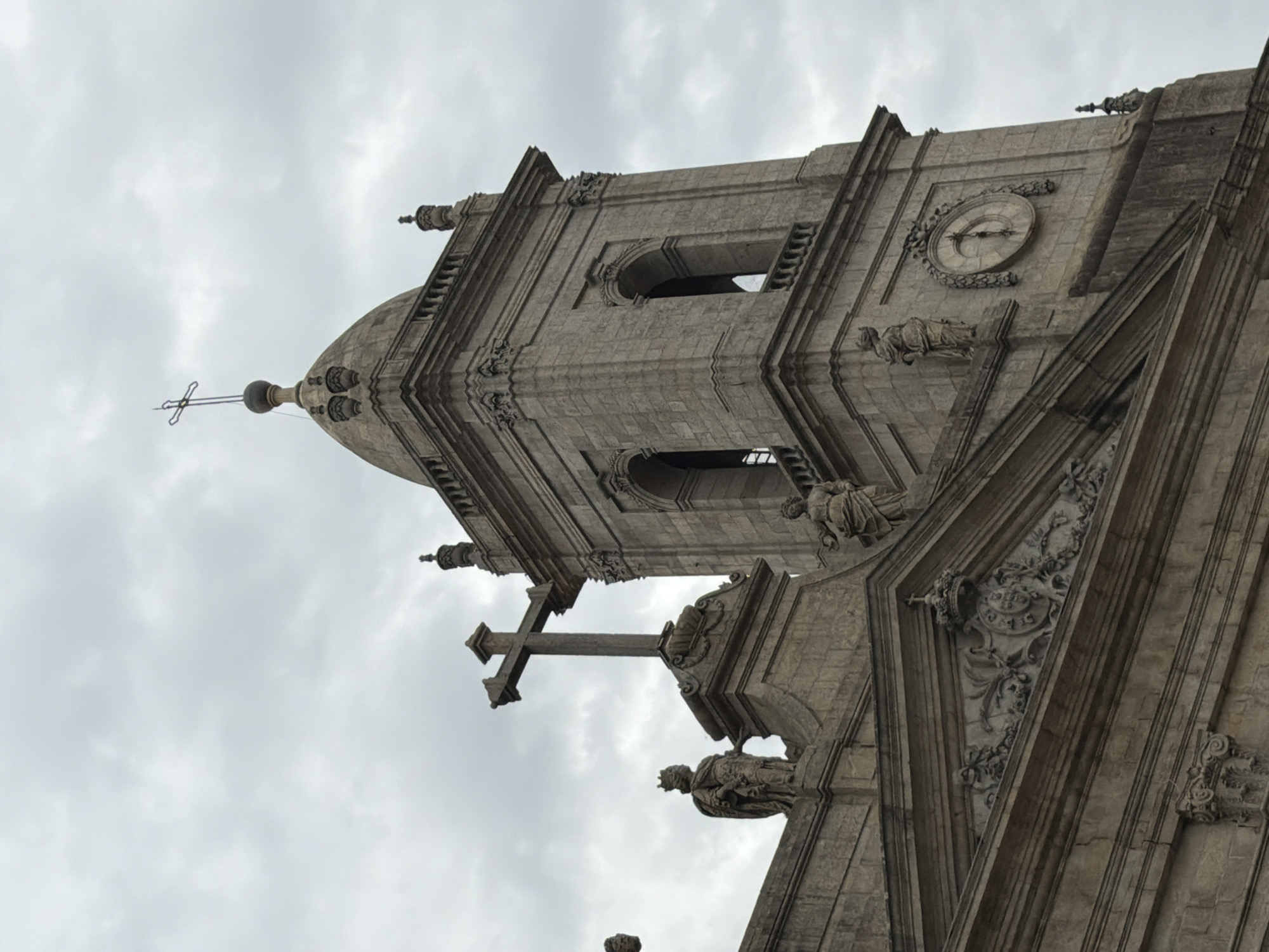 This is a close-up, low-angle shot of the exterior of a large, stone building, likely a cathedral or church, with intricate architectural details. The building's facade is made of grey stone, carved with religious iconography and decorative elements. Prominently featured are a large clock on the facade, a cross atop a structure, and several sculpted figures of people in historical attire, possibly saints or royalty, adorning ledges and niches. The style of the architecture suggests it is of historical significance, likely European. The sky above is overcast and cloudy, indicating a possibly cool or humid day. The image is rotated 90 degrees counter-clockwise, with the sky occupying the left side of the frame and the building's architecture on the right. The lighting is diffused, typical of an overcast day. There is no visible text in the image. The overall impression is one of grandeur and historical weight, with the cloudy sky adding a somber or dramatic mood.