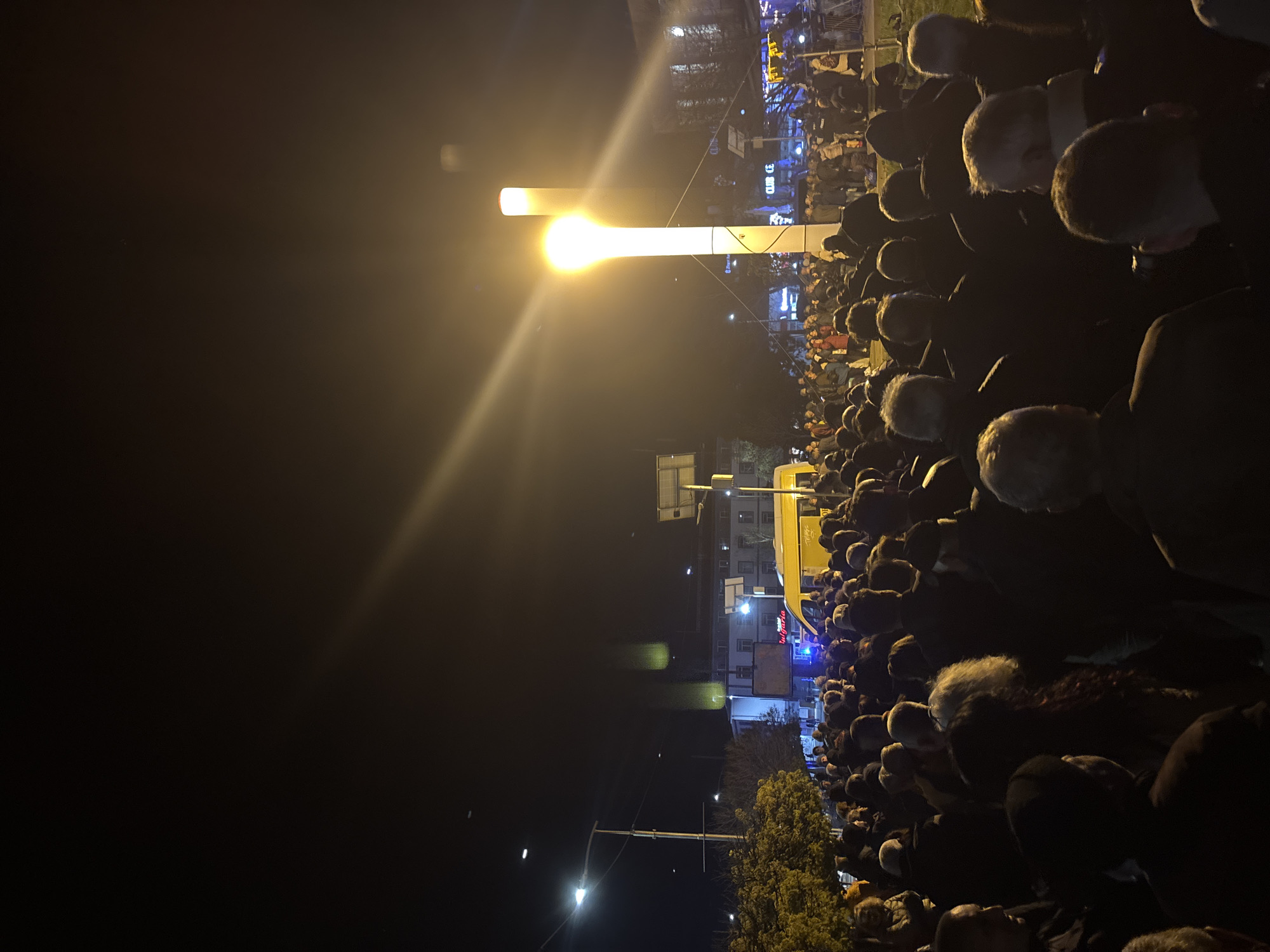 A large crowd of people is gathered at night in Kardzhali, Bulgaria, under a bright light post and illuminated buildings. The majority of people are facing towards the light, suggesting they are watching or attending something. The setting appears to be urban, with trees and streetlights visible as well. A vehicle, possibly a van, is present among the crowd. The people's heads are visible, with a variety of dark and gray hair colors.