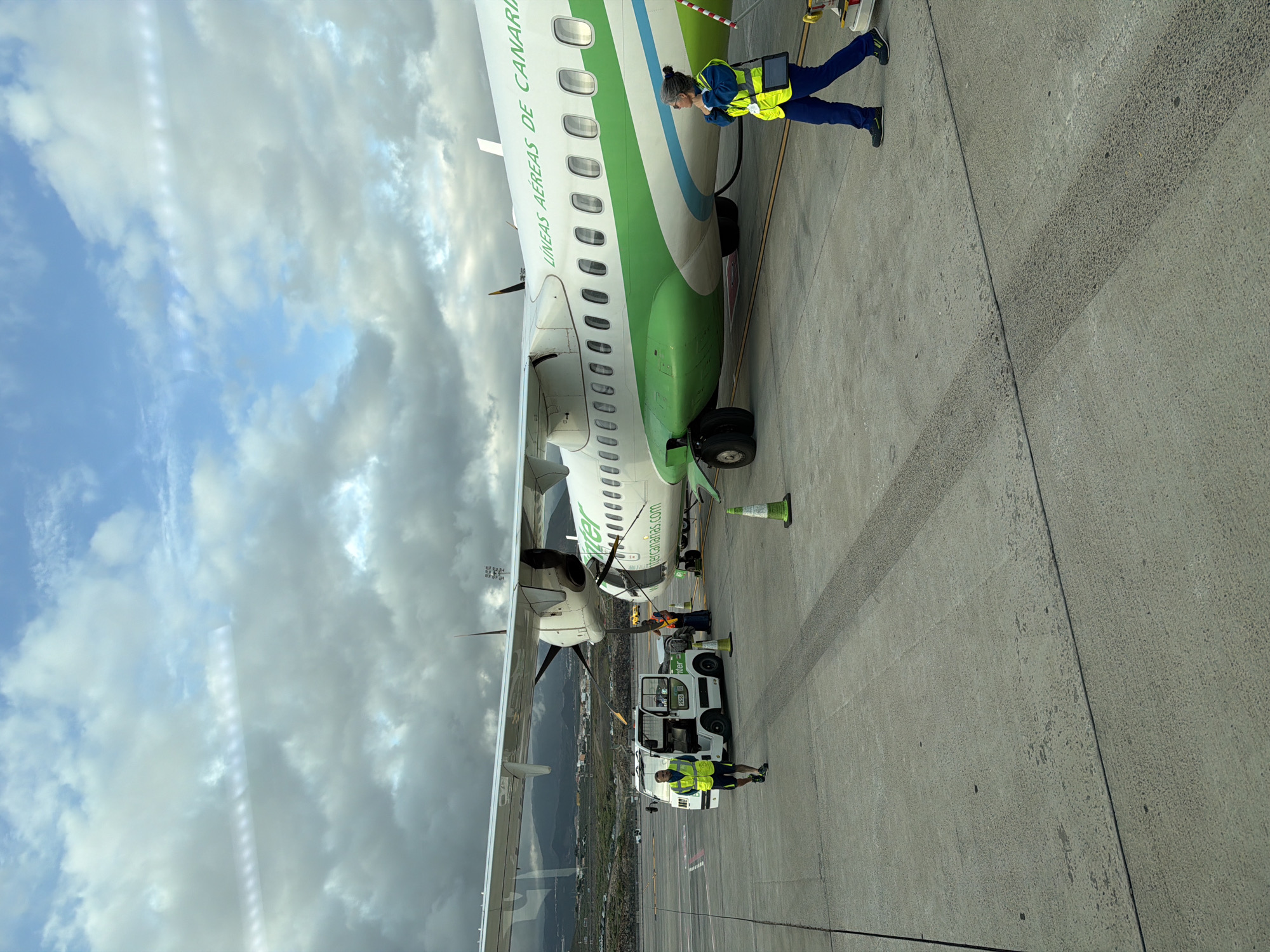 An active airport tarmac scene unfolds during the daytime under a partly cloudy sky, featuring an aircraft and ground personnel. The main subject is a turboprop aircraft, distinctively painted with a white fuselage, a prominent green stripe along its lower half, and a thinner blue stripe above it. The airline's name, "BINTER LÍNEAS AÉREAS DE CANARIAS," is visible along the top of the fuselage, while "bintercanarias.com" is printed below the windows.
Two ground crew members, both wearing high-visibility yellow vests and dark blue pants, are attending to the aircraft. One female crew member is positioned near the front of the plane, intently looking down at a tablet or device she holds in her hands, suggesting a pre-flight or post-flight check. Further towards the rear of the aircraft, another male crew member stands beside a white ground support vehicle, which has "inter" written on its side (likely part of "Binter"). An orange traffic cone is placed near the front landing gear of the plane.
The ground is a grey concrete tarmac, showing some tire marks or wear. In the background, beyond the plane's wing and tail, a sprawling landscape of mountains or hills is visible, dotted with buildings, under a mix of blue sky and scattered white and grey clouds. The overall impression is one of routine airport operations, likely in the Canary Islands, given the airline's name and the suggested location of Granadilla de Abona, Spain.
