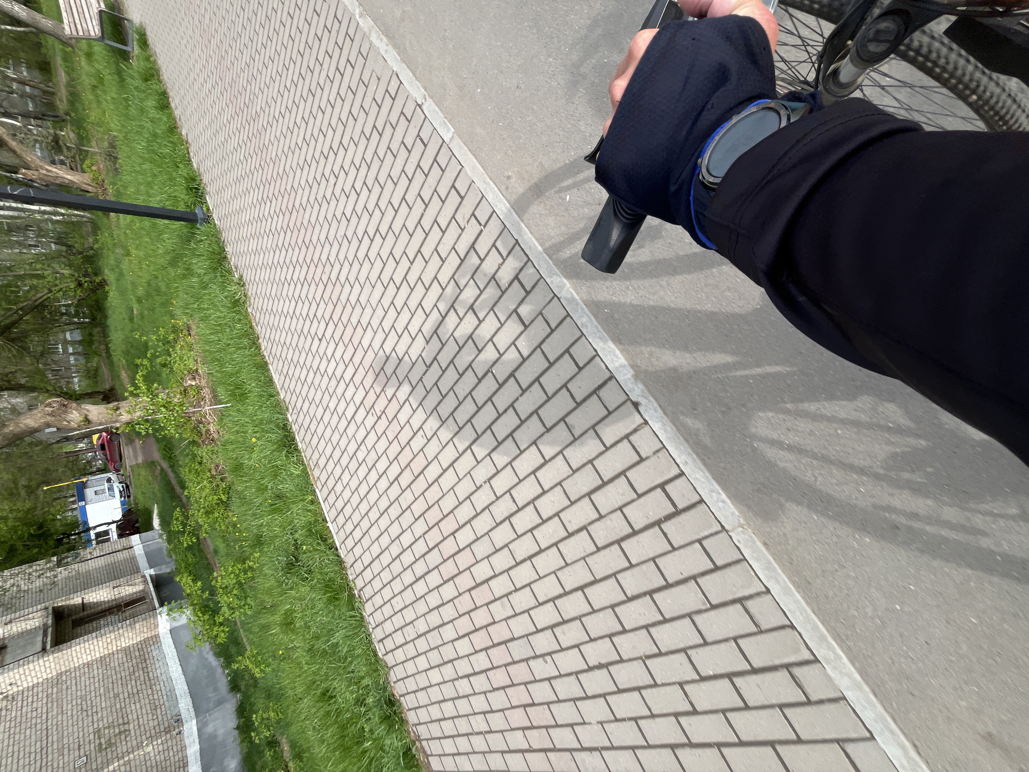 The image presents a first-person perspective while riding a bicycle. A hand covered in a black glove, with a smartwatch on the wrist, grips the bicycle handlebar. The bicycle's front wheel is partially visible. The foreground features a paved path consisting of rectangular gray bricks. Adjacent to the path is a strip of green grass and sparse vegetation. In the blurred background, a building and a vehicle can be discerned.
Based on the context provided, it is reasonable to assume that this scene is located in the city of Chaikovsky, Russia.