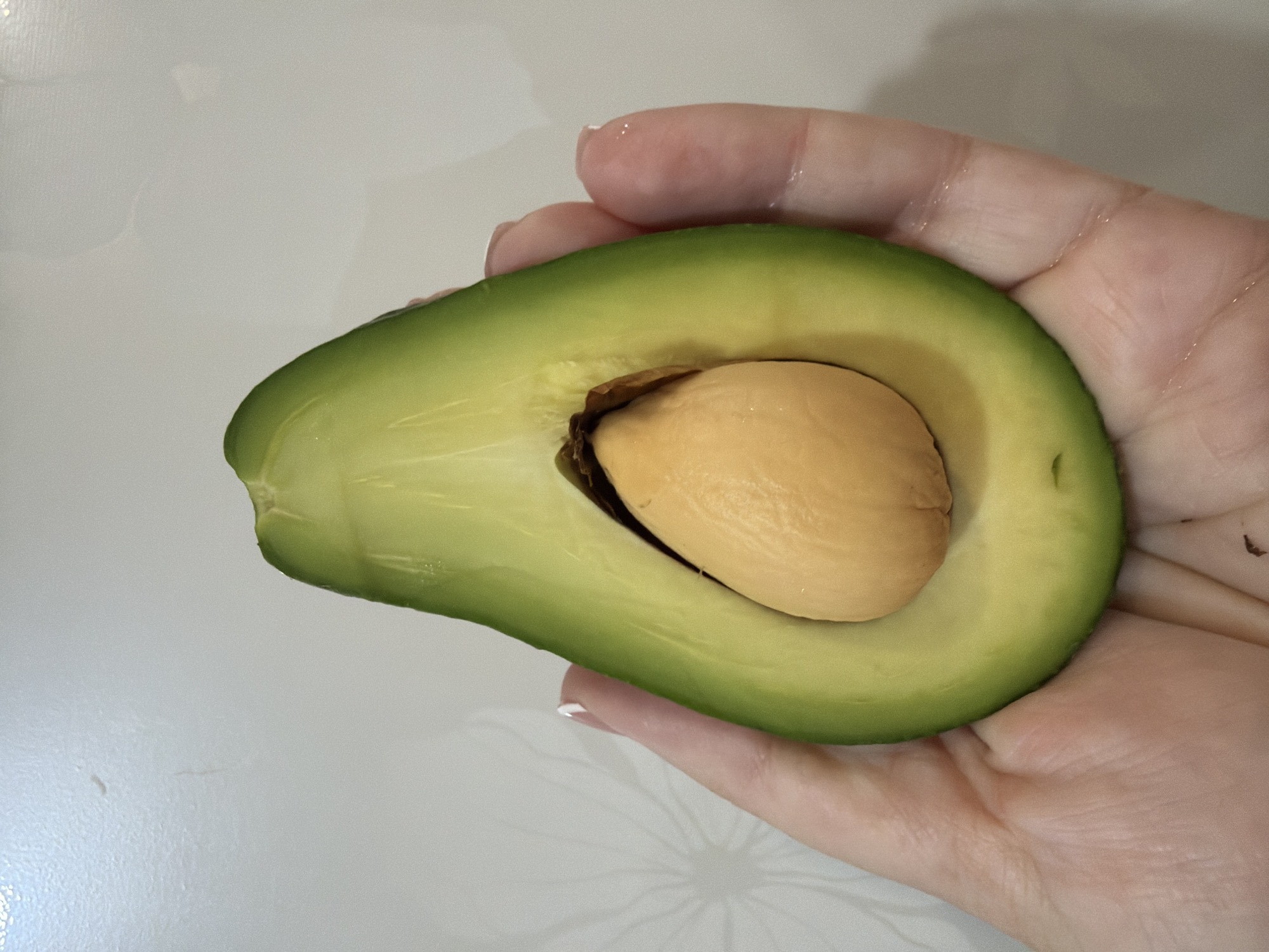 A medium close-up captures a human hand holding half of an avocado. The avocado, cut lengthwise, features a smooth, green outer layer and a lighter, yellowish-green interior. Its seed remains intact. The hand holding the avocado is fair-skinned, with manicured fingernails bearing a white polish. The background is a light, reflective surface. No details indicate the location being Chaikovsky, Russia.