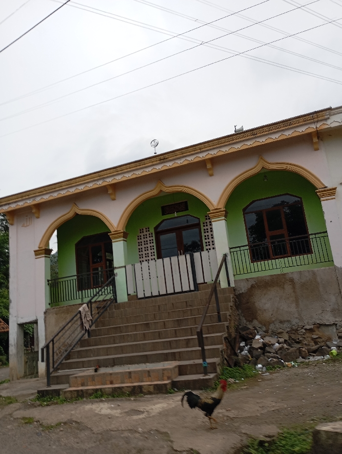 The image shows a two-story building with a concrete staircase leading up to a set of double doors. The building is painted white with a yellow trim, and has a green painted wall below the windows. The building has a series of arches in the facade, with some of the windows open. The building has a large, gray concrete wall under it. There are power lines running overhead. A brown and white rooster is walking at the bottom of the stairs. The building appears to be a mosque. The location is at the coordinates (latitude: -6.971, longitude: 107.519) in Kecamatan Kutawaringin, Indonesia.