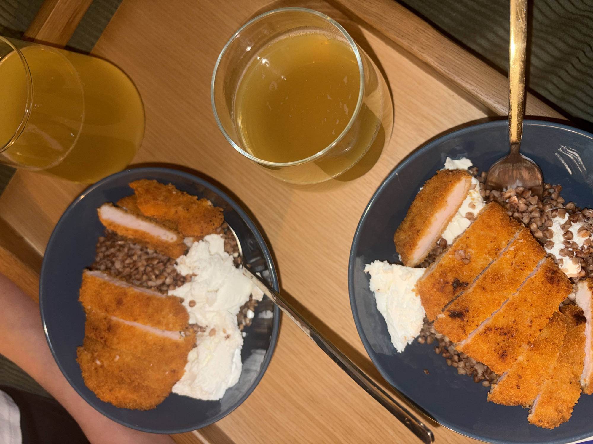 Two plates of food and two glasses of orange liquid are arranged on a wooden surface. The plates are dark blue and contain buckwheat, mashed potatoes, and sliced breaded cutlets. A golden spoon rests on the left plate, and a silver fork is on the right plate. The glasses are clear and filled with an orange beverage. The scene appears to be an indoor setting, possibly a home or restaurant, in Tel-Aviv, Israel.