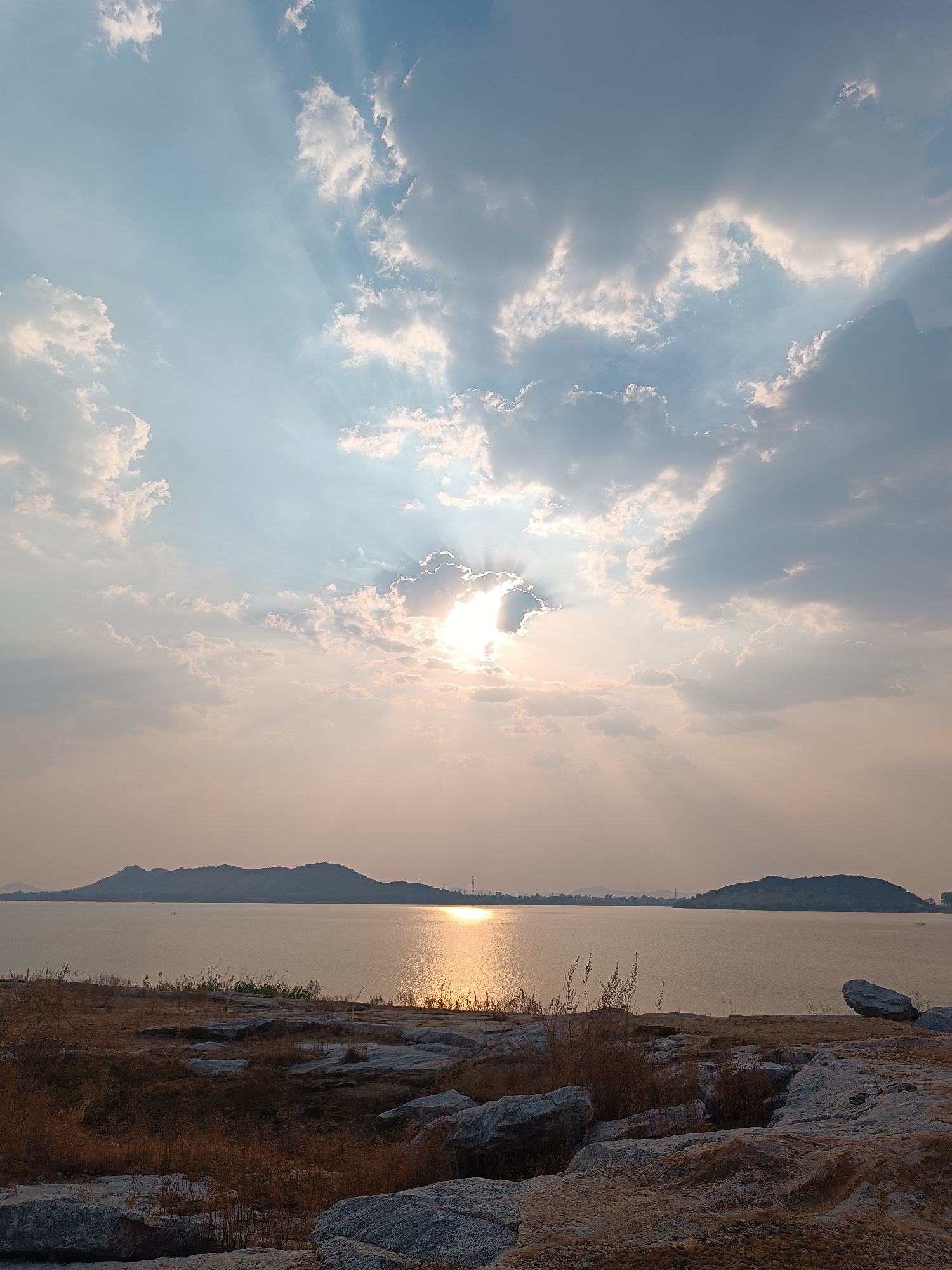 A landscape photograph captures a body of water under a sky with scattered clouds and the sun shining through them. The sun's rays are visible, creating a diffused light effect. In the distance, there are two land masses, possibly islands or hills, on the horizon line of the water. The foreground consists of rocky terrain with some vegetation. The scene suggests a tranquil, natural environment, possibly a lake or coastal area at sunset or sunrise. The location is in the city of Unknown, Unknown.