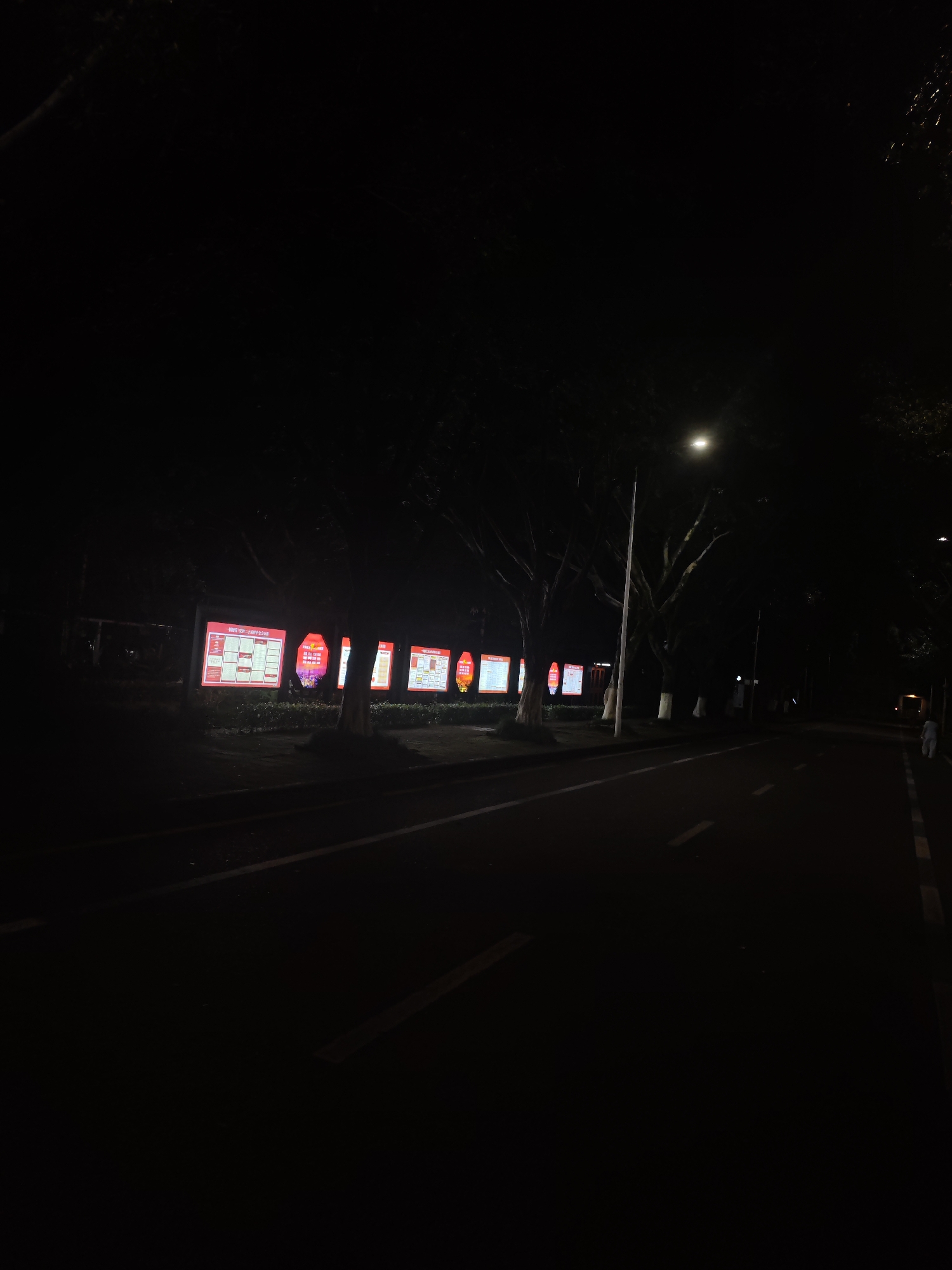 The image depicts a street scene at night in the Chongqing High-tech Zone, China. A paved road with white lane markings is visible in the foreground, leading into the distance. On the left side of the road, a row of illuminated billboards or display screens is set up. These screens, predominantly red and white, display various information, including charts, graphs, and text, likely advertisements or public notices. Several trees with visible trunks and branches line the sidewalk beside the billboards, their leaves silhouetted against the dark sky. A street lamp with a bright light is visible above the trees, illuminating a portion of the scene. In the distance, a lone figure, appearing to be a person, is walking on the right side of the road, their silhouette distinct against the dim light. The overall atmosphere is dark and quiet, characteristic of a nighttime urban environment. The illuminated displays are the primary source of light apart from the street lamp, creating a stark contrast with the surrounding darkness.