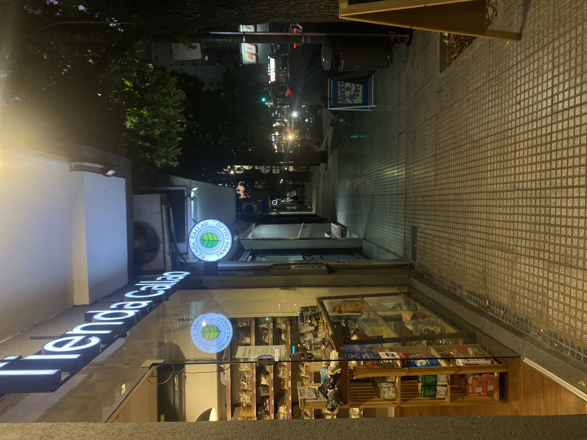 This nighttime, street-level photograph depicts a commercial sidewalk and street scene, identified as being on or near Callao Avenue in Buenos Aires, Argentina, due to the prominent "Tienda Callao" signage. On the left, the storefront of "Tienda Callao" is illuminated by its white channel letters and a circular sign displaying a green leaf logo. Through the large glass window, the store's interior is visible, revealing shelves stocked with various packaged food items and jars, along with a wooden display unit. The sidewalk is paved with light-colored, square-patterned tiles and extends into the distance. A black public trash bin and a partially visible blue A-frame sign, reading "ABIERTA MERC" (Open Market), are situated on the sidewalk. A large tree with a dark trunk and green foliage stands to the right, adjacent to the street. The street itself shows parked vehicles and active traffic, with oncoming car headlights visible in the mid-ground. Distant streetlights and building signs contribute to the overall urban illumination. One pedestrian is discernible walking away on the sidewalk further down the street. The scene is observed under clear conditions.
