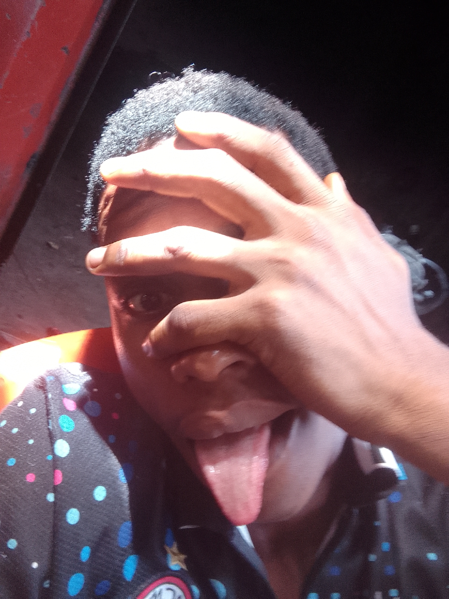 A person with dark skin and short, dark hair is pictured indoors with a hand obscuring the upper portion of their face. One eye is visible, peering between the fingers. The person is sticking their tongue out. They are wearing a dark shirt with colorful polka dots. A red surface is visible on the left side of the frame, and headphones are around the neck on the right. The background is dark. There is no information given to indicate location or city.