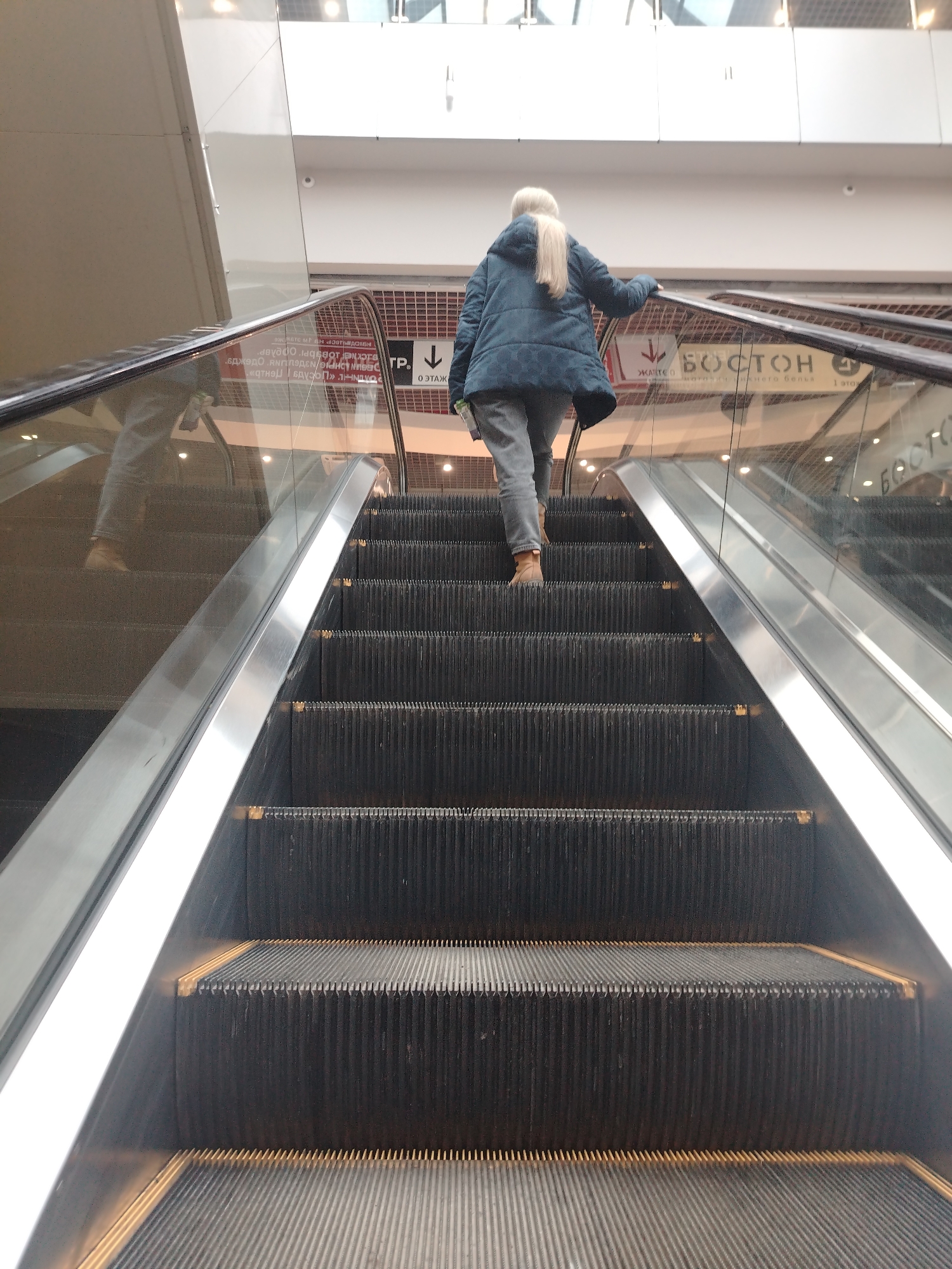 A woman with long, blonde hair is riding up an escalator in what appears to be a shopping mall in Izhevsk, Russia. She is wearing a dark blue puffer coat, gray pants, and brown boots. The escalator has a stainless steel handrail and dark gray steps. Signs with text in Cyrillic script, including the name "BOSTON," are visible on the glass panels adjacent to the escalator. The escalator is heading upwards. The woman is holding onto the handrail. The environment appears to be a modern, brightly lit shopping center.