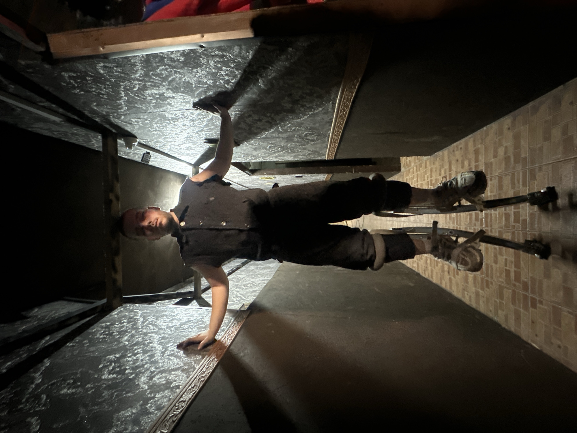 A single male individual stands centrally in a narrow, dimly lit indoor corridor. He faces forward, looking directly at the camera, with his arms extended horizontally, pressing his palms against the adjacent walls for stability. The individual wears a dark, short-sleeved collared shirt, dark trousers, and light-colored athletic shoes. He is elevated on a pair of silver-colored stilts, secured to his lower legs and feet.
The corridor walls feature distinct sections: the upper portion is covered with dark wallpaper displaying a light-colored, intricate ornamental pattern, while the lower portion is a solid dark color. A decorative molding with a repeating, possibly gilded or metallic, pattern separates these two wall sections. The floor is composed of small, rectangular, light brown tiles arranged in a grid pattern. The ceiling is dark, revealing visible structural elements. Illumination originates from above and slightly in front of the subject, casting shadows behind him and highlighting the wall textures. The corridor extends into the background, with a faint doorway visible on the left and a red object or fabric partially visible on the far right. The specific location within the city of Saratov, Russia, cannot be determined from the provided image.
