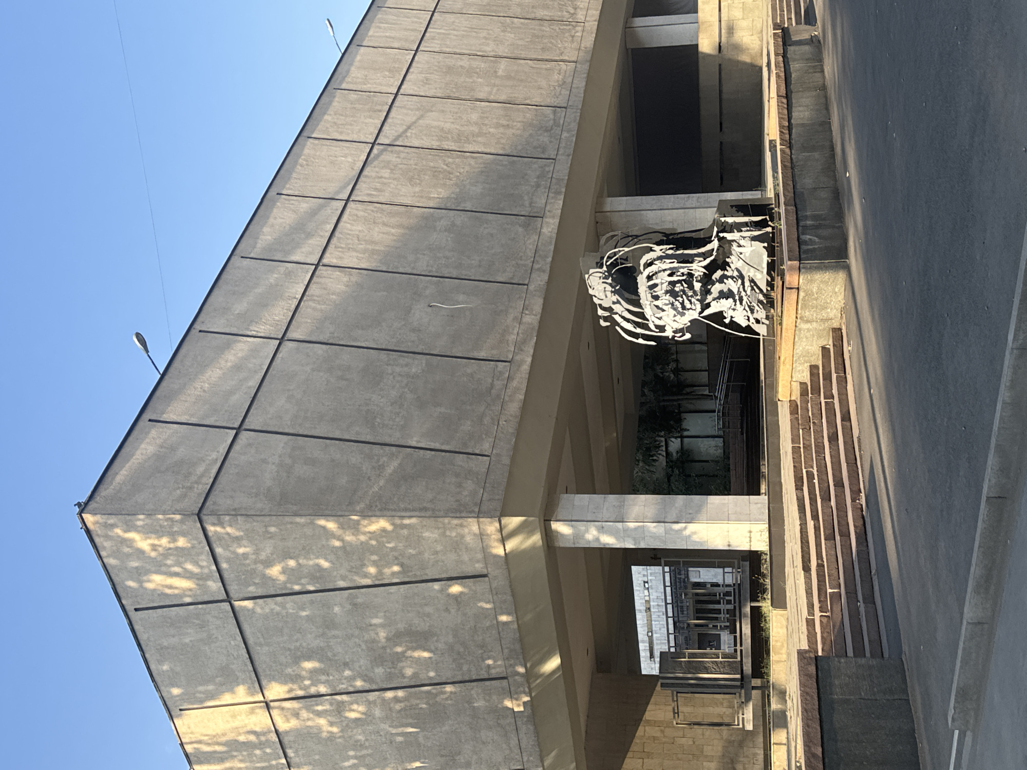 The image shows an outdoor scene in front of a brutalist building, possibly a museum or government building, in Bishkek, Kyrgyzstan. The building is made of concrete and features a distinct geometric design with angled panels and vertical dark lines. In the foreground, there is a modern abstract sculpture of a head, rendered in a metallic, wireframe style. The sculpture is placed on a concrete pedestal surrounded by red stone. To the left of the sculpture, a wide set of stone stairs leads up to the building's entrance. Sunlight casts long shadows across the scene, indicating it is likely late afternoon or early morning. The sky is clear and blue. The overall impression is one of civic architecture and public art.