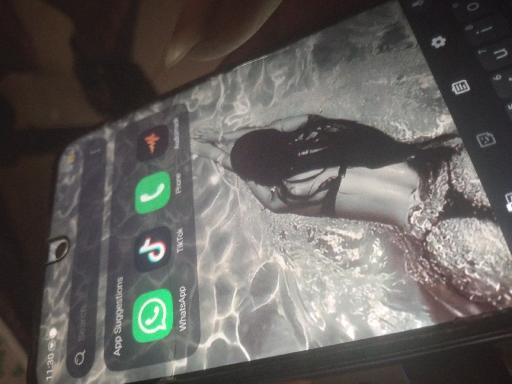 The image shows a close-up of a smartphone screen displaying a wallpaper of a person submerged in water. The wallpaper is a grayscale image of a person with long dark hair, appearing to be swimming or posing in water with sunlight rippling on the surface. The phone's interface is visible, with app suggestions including WhatsApp, TikTok, Phone, and Audiomack. The top left corner of the screen shows the time as 11:30 and a battery icon. The phone itself is held at an angle, and a person's hand is partially visible on the right side of the frame, holding the device. The overall scene appears to be indoors, with a dark background beyond the phone's screen. There is no information within the image that indicates a specific location in the city of Durimi, Nigeria.
