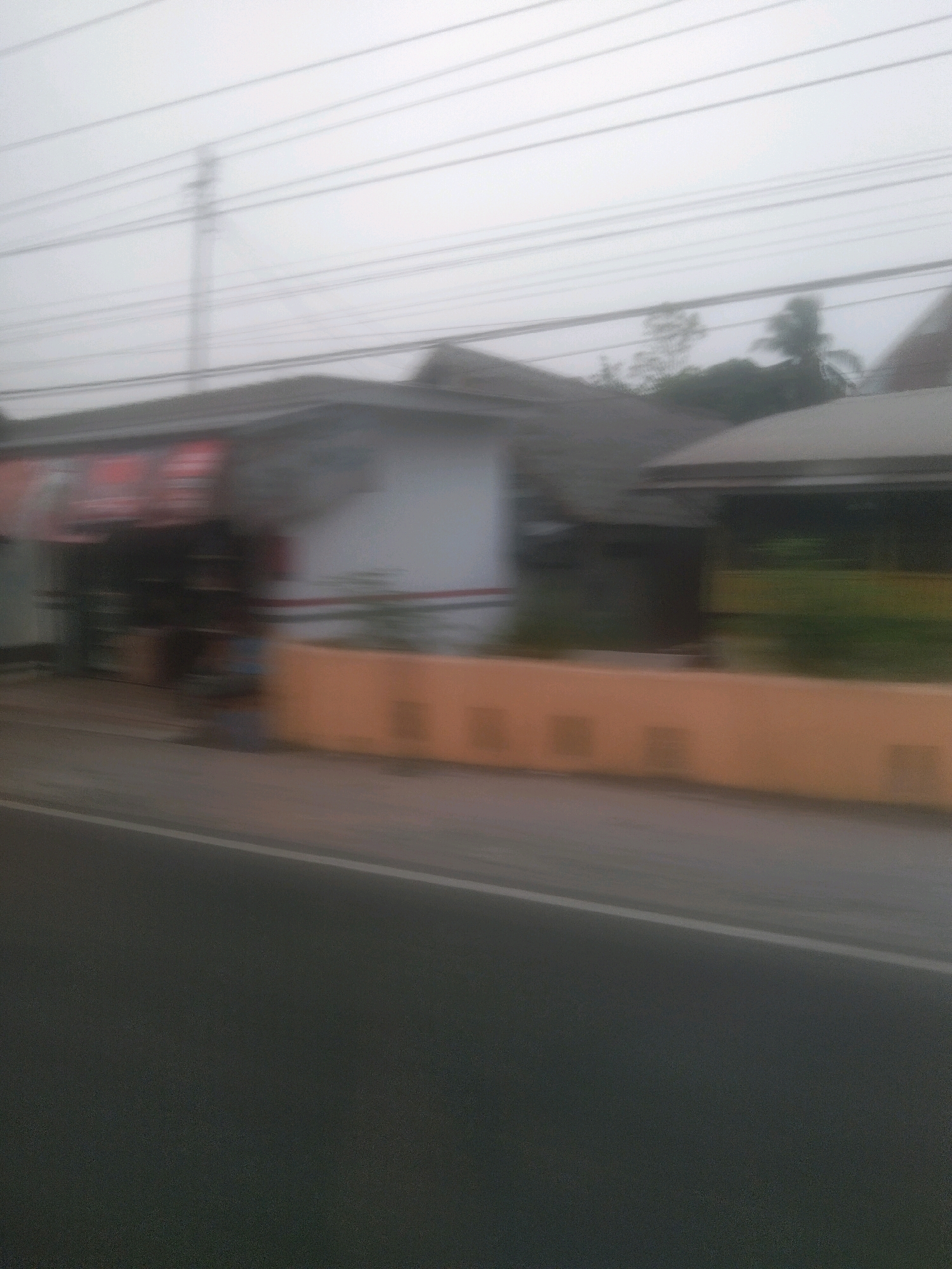 The image depicts a blurred street-level view of buildings and a road, likely captured from a moving vehicle. In the foreground, a dark asphalt road with a white line is visible. Behind the road, a low, orange barrier or wall runs horizontally across the scene. Beyond the barrier, several buildings are visible, some with sloping roofs and white walls. A portion of a building on the left has red signage that is too blurred to read. Trees, including what appears to be a palm tree, are visible behind the buildings, silhouetted against a hazy sky. Overhead, multiple power lines are strung across the frame. The overall impression is one of a somewhat overcast day in a developed, possibly commercial, area. The location is identified as Kayu Agung, Indonesia.