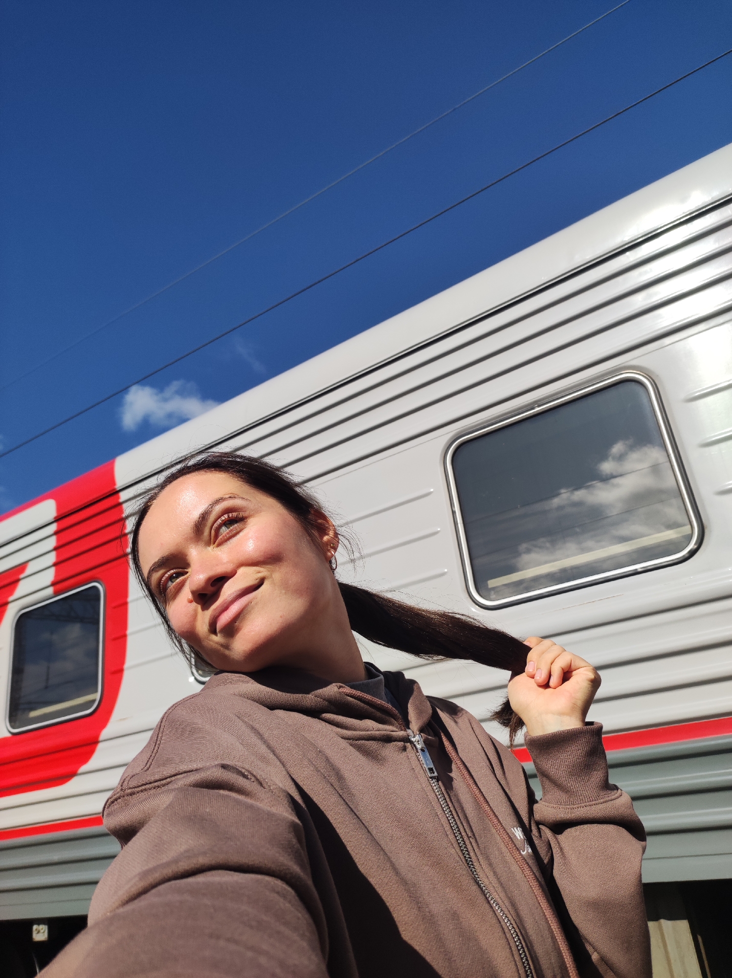 A young woman with dark hair pulled into a ponytail, wearing a brown zip-up hoodie, poses for a selfie outdoors. She is looking upwards and to her left, with a slight smile. Behind her, a train car with red and white stripes and horizontal grey paneling is visible. The sky above is a clear blue with a few wispy clouds. The train car is likely located at a train station in Kirov, Russia, given the context.