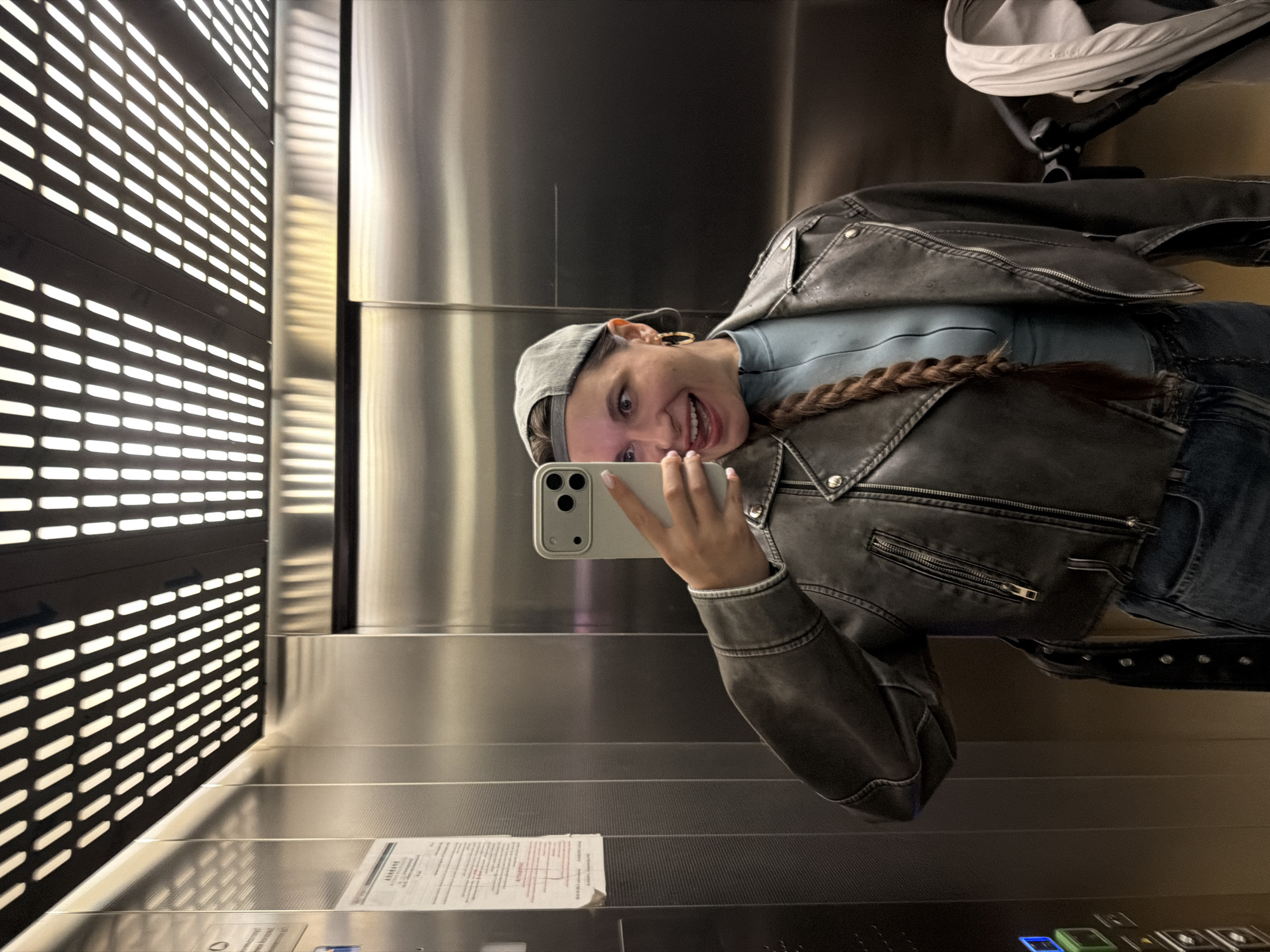An individual is taking a selfie in an elevator. The person is wearing a grey baseball cap, a grey leather jacket, and a blue turtleneck. They have a long braid of brown hair. The elevator has stainless steel walls and a ceiling with rows of lights. There is a document posted on the left wall of the elevator. The individual is sticking out their tongue and making a playful expression. A stroller is visible to the right of the person.