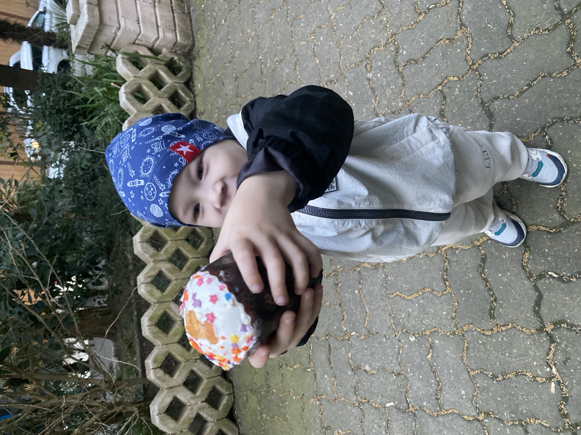 A young child stands outdoors on a paved surface, holding a decorated pastry. The child wears a blue patterned hat, a white and black jacket, and light-colored pants with blue and white sneakers. The pastry appears to be a dark brown color with a white frosting covered in colorful sprinkles. The paving is made of gray interlocking blocks with small debris between them. Background elements include greenery, concrete blocks, and a glimpse of a white vehicle. The location of the city of Sochi, Russia, is not provided in the image.