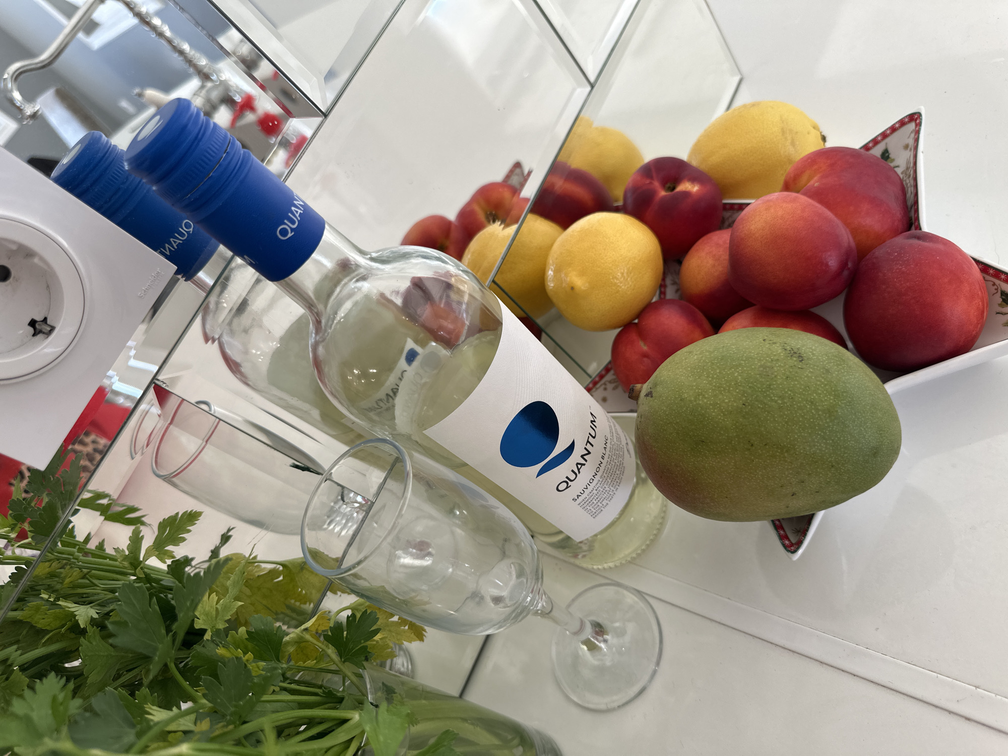 The scene is set indoors in Asenovgrad, Bulgaria, depicting a still life arrangement on a glossy white surface against a mirrored wall. To the left, a clear glass vase contains a bundle of fresh green herbs, likely parsley or celery leaves. An empty stemmed wine glass stands nearby. In the center, a bottle of 'QUANTUM Sauvignon Blanc' white wine, identifiable by its blue screw cap and white label with a blue logo, rests on the surface. To the right, a decorative white bowl with a red and green pattern on its rim holds an assortment of fruits including several red-orange nectarines or peaches and yellow lemons. A large green mango with reddish blush is positioned in the foreground, separate from the bowl. The mirrored wall behind reflects the wine bottle, the empty glass, and the fruits. In the upper reflected area, a white electrical wall outlet with 'Schneider' branding is visible, along with the reversed 'QUANTUM' text from the wine bottle's blue cap.
