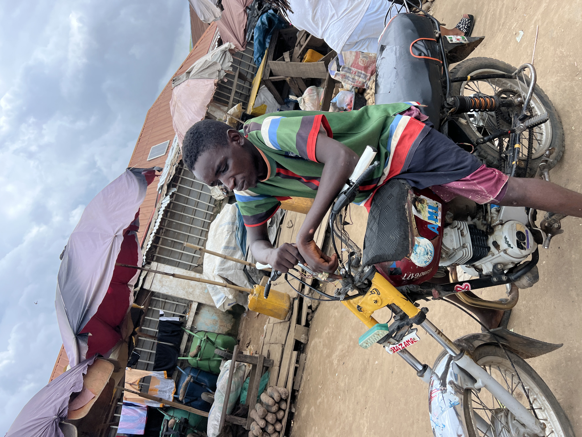 A young person sits on a red and black motorcycle at a market in Asaba, Nigeria. The individual is wearing a green, red, blue, and white striped t-shirt and purple shorts. Their hands are on the handlebars of the motorcycle, which has a yellow handlebar support. The background shows market stalls with various goods, including textiles hanging on a line, produce, and sacks of goods. Several umbrellas are set up to provide shade. The ground is dirt.