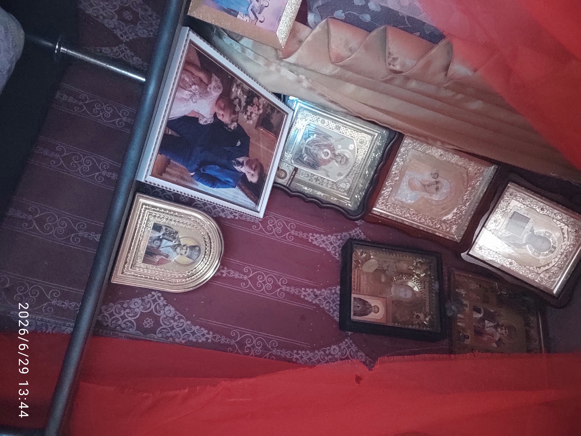 A religious collection of icons and framed photographs are displayed in a room, possibly in Savyntsi, Ukraine. The collection is arranged on a shelf or ledge against a wall decorated with a dark red, patterned wallpaper. Red fabric drapes down from the top left and right of the frame, obscuring parts of the background.
The displayed items include several framed religious icons depicting saints, with ornate gold or silver borders. One prominent icon in the center shows a figure of a woman with a child. There is also a framed photograph of a man in a blue suit and a young girl in a white dress. Another framed photograph on the far left appears to show a group of people. A small, arched icon with a depiction of a bearded man in religious attire is visible in the lower right corner.
The overall impression is of a domestic altar or devotional space. The lighting appears to be dim, with some reflections on the glass of the frames. The date stamp "2026/6/29 13:44" is present in the lower right corner.
