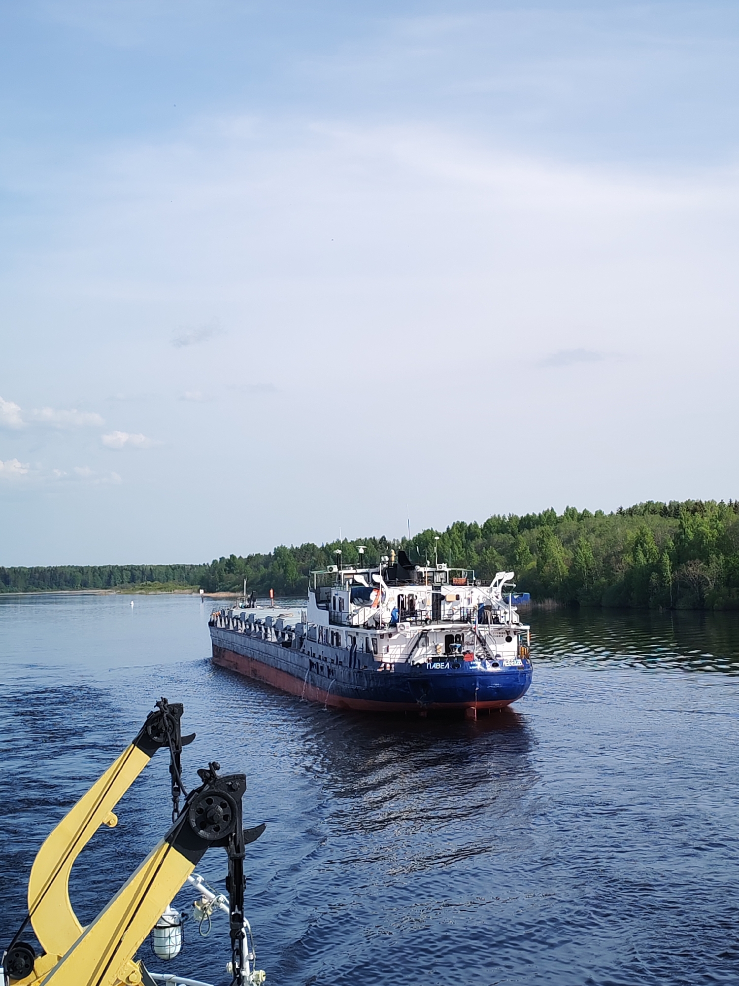 The image shows a barge on a wide body of water, likely a river, with forested banks in the background. The barge is painted with a blue hull and red waterline, topped with a gray deck and white superstructure. Text on the stern indicates it's named "PAVEL LEBEDEV." Yellow and black davits are visible in the foreground, suggesting the photograph was taken from another vessel. The sky is partially cloudy. There's no specific information to confirm the location as Edoma, Russia, but the river landscape is consistent with such regions. No people are clearly visible.