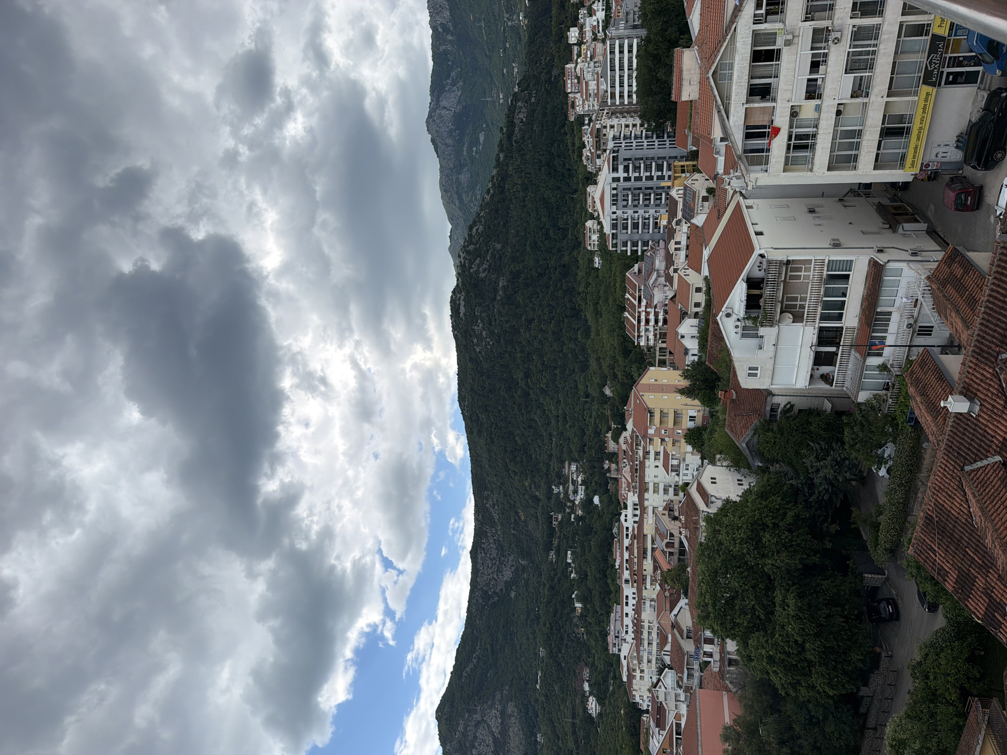 Here's a detailed description of the image:
The image showcases a cityscape nestled against a mountain range, likely captured from an elevated vantage point in Budva, Montenegro. The sky is partially overcast with a mix of heavy, dark clouds and patches of bright blue sky peeking through.
A prominent, steep mountain is covered in dense green vegetation. Along the base and slopes of the mountain, there's a cluster of buildings of varying heights and architectural styles. The buildings predominantly feature white facades and red-tiled roofs, a characteristic of Mediterranean architecture.
Closer to the foreground, more buildings are visible, including taller apartment blocks, interspersed with smaller residential homes. Some structures have visible balconies and windows. Vegetation, including trees and shrubs, dots the landscape, adding to the natural element of the urban setting.
On the right edge of the frame, a street can be seen with a few parked cars. A sign with yellow background and black writing hangs by the street and might indicate a business. Red flags are visible on balconies.
The composition suggests a blend of natural mountainous terrain and urban development. The lighting is somewhat diffused due to the cloud cover.