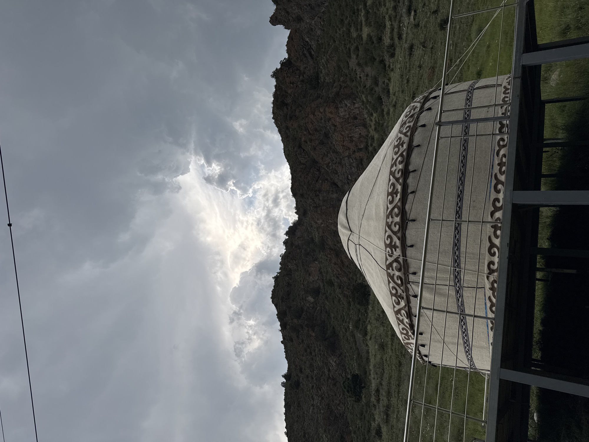 This is a medium shot of a yurt situated on a raised platform with a metal railing in front of a steep, rocky hillside. The yurt is decorated with intricate brown and beige patterns around its circumference and a decorative band around its upper section. The hillside behind the yurt is covered in sparse green vegetation and dotted with trees. The sky above is filled with dramatic, heavy clouds, with a patch of bright sunlight breaking through near the center. A thin power line stretches horizontally across the top of the frame. The scene appears to be in a natural, possibly mountainous, outdoor environment, consistent with the location of Kyzyl-Korgon, Kyrgyzstan. There are no people or animals visible in the image.