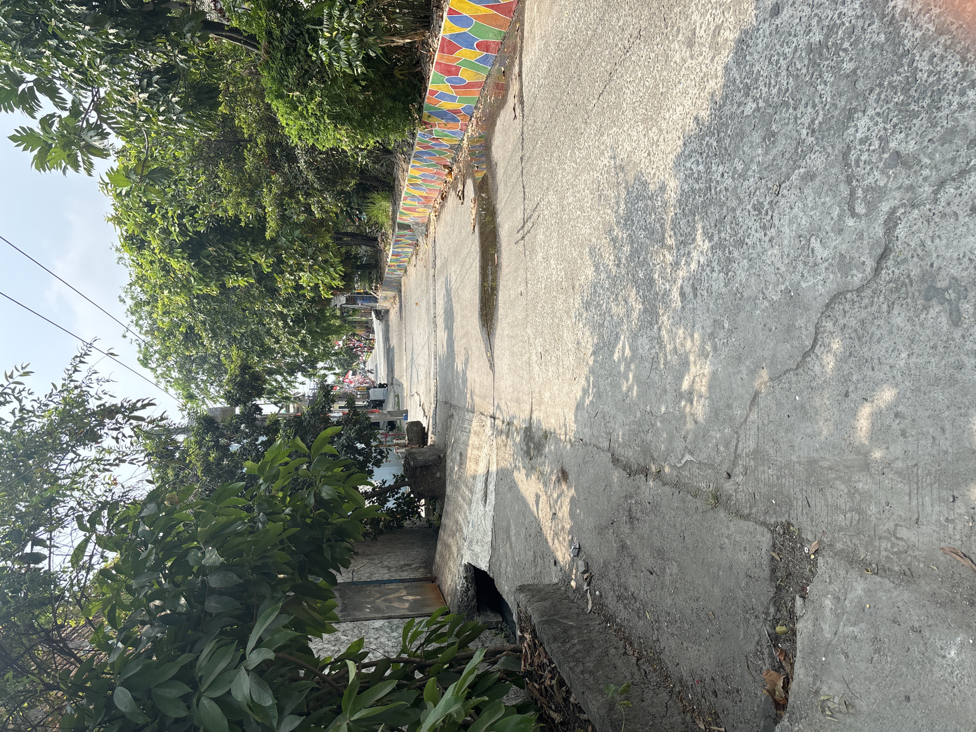 The image depicts a narrow, paved street in Telagamurni, Indonesia, lined with mature trees and residential buildings. A concrete drainage channel runs along the left side of the street, and a section of the pavement is cracked and shows signs of wear. On the right side, a low, vibrantly painted wall with a geometric pattern of red, yellow, green, and blue rhombuses and circles borders the street. Puddles of water reflect the colorful wall and the sky. The street recedes into the distance, where more trees and buildings are visible, along with a few people and motorcycles. Overhead, power lines are strung across the clear, bright sky. The scene appears to be a residential or local commercial area, bathed in bright sunlight.