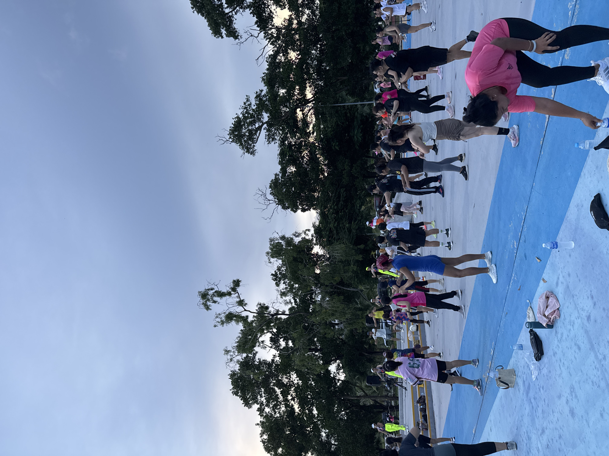 The image depicts a group of people engaged in an outdoor fitness class, possibly a workout or exercise session, at what appears to be a public park or recreational area in Phuket City Municipality, Thailand. The setting features a blue and white tiled ground, bordered by a yellow line, with lush green trees providing a backdrop. The sky is overcast, suggesting either early morning or late afternoon, with the low light casting soft shadows.
The individuals, predominantly women, are dressed in athletic wear in various colors, including pink, black, white, yellow, and blue. They are positioned in a line, performing exercises that involve stretching and core strengthening, such as planks and lunges. Some are holding water bottles, indicating a sustained activity. The overall mood seems focused and energetic, with participants concentrated on their movements. The presence of trees, the specific tiling of the ground, and the likely warm climate implied by the attire suggest a tropical or subtropical location, consistent with Phuket. No visible text is present in the image.