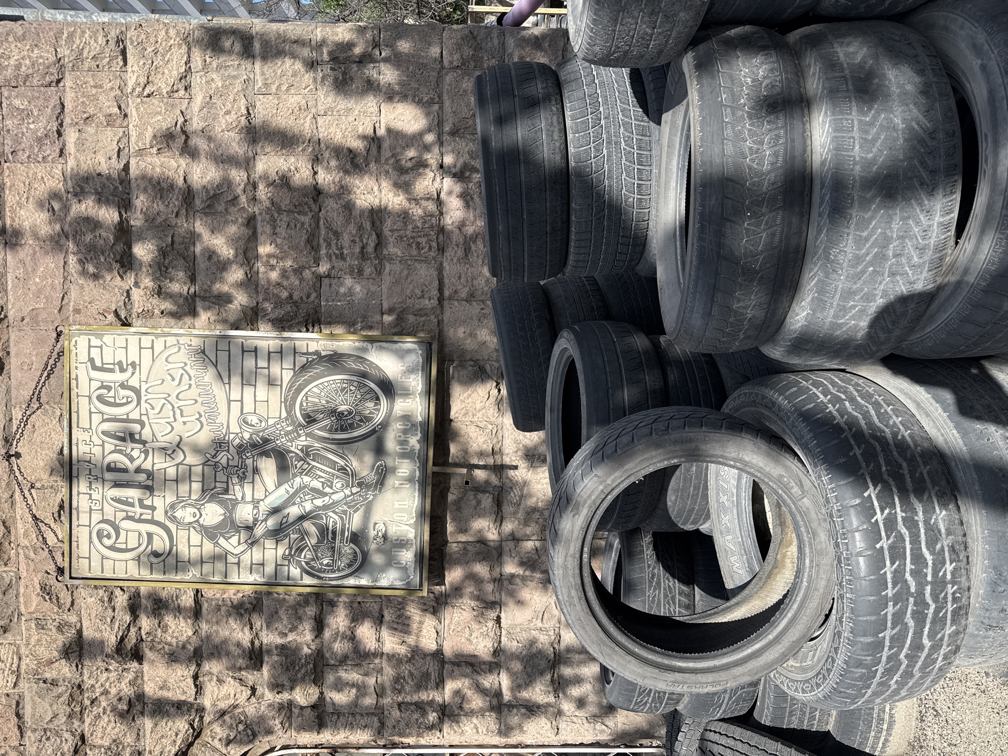 The image depicts a scene at a garage in Yerevan, Armenia. A sign with "GARAGE" written on it and an illustration of a woman and motorcycle hangs on a stone wall. Adjacent to the sign is a stack of used tires. The environment appears to be outdoors, with natural light and tree shadows cast on the wall. There are no people visible in the image.