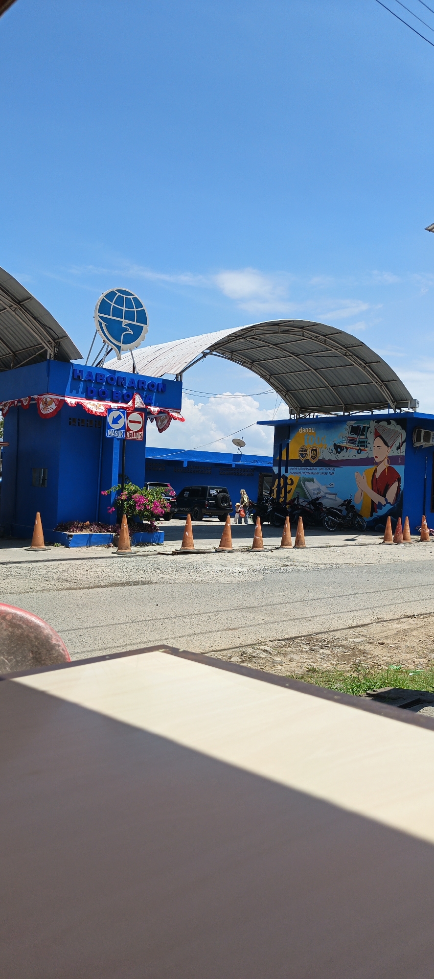 The image captures a scene in Tigaras, Indonesia, featuring a bright blue building serving as an entrance gate. Above the entrance, a prominent logo with a globe and an eagle is visible. The building is adorned with red and white striped banners. Several orange traffic cones are strategically placed along the road in front of the gate, directing traffic flow.
To the right of the entrance, a mural is painted on the side of a building, depicting a person in traditional attire, a boat, and what appears to be a mountain landscape. Several motorcycles are parked in front of this building, and a black SUV is parked further down the road.
The environment suggests a sunny day, with a clear blue sky and scattered clouds. The foreground shows a blurred surface, possibly a table or a car hood, casting a shadow. The overall scene appears to be a transportation hub or border crossing point.