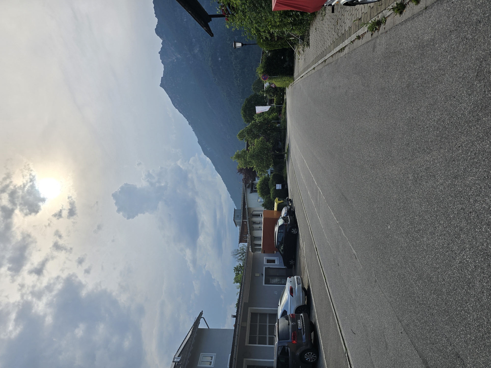 The image depicts a residential street in Garmisch-Partenkirchen, Germany, with a large mountain range dominating the background. On the left side of the street, a gray building with a garage door is visible, with several cars parked in front of it. The cars are a dark gray SUV, a white sedan, and a black hatchback. The street is asphalt, with a paved sidewalk on the right side. A lamppost stands on the sidewalk next to a lush green hedge, and further down, a red tarp covers an object, possibly a trailer or equipment. The sky is partly cloudy with the sun peeking through, casting a bright glow. The overall scene suggests a peaceful, suburban environment with a strong natural backdrop.
