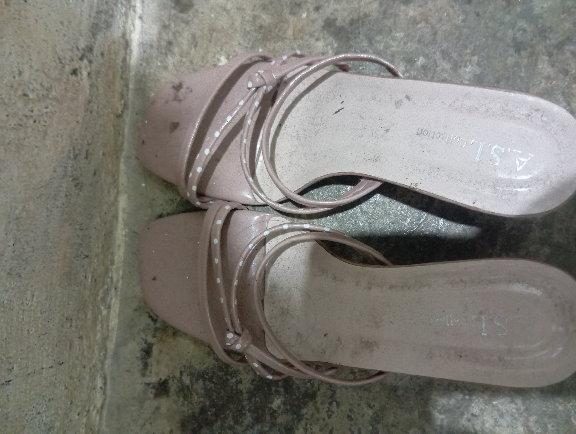 A pair of dusty, light brown flat sandals with thin, strappy uppers decorated with white polka dots are placed side-by-side on a rough, grey concrete floor in what appears to be an outdoor or semi-outdoor space. The sandals have a textured sole with the embossed text "A.S.L. Collection" visible on the right sandal. The background consists of a textured, possibly concrete or stone wall with some darker stains and a horizontal crack. The lighting is dim, casting subtle shadows. This scene is located in Rejosari, Indonesia.