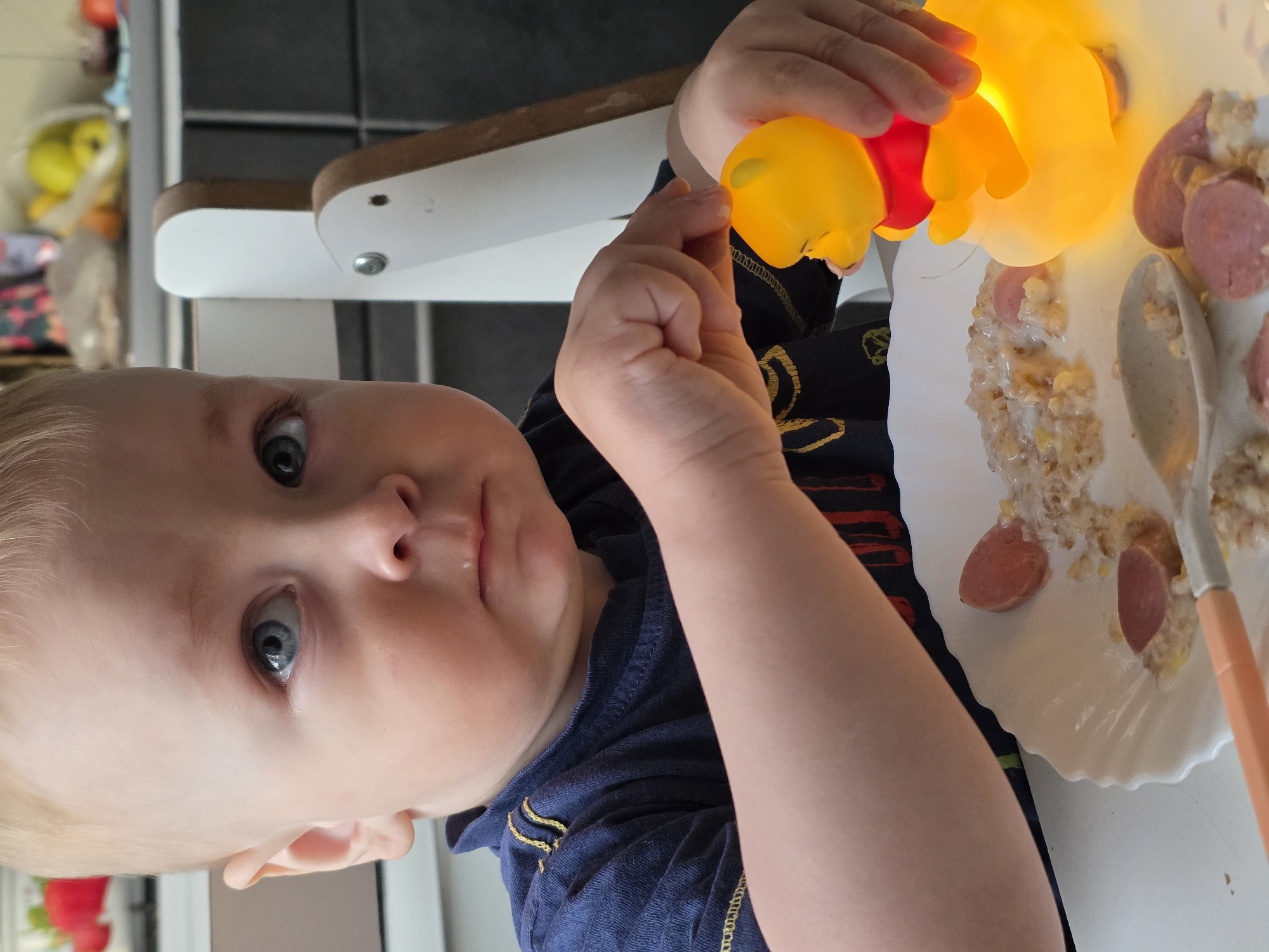 A baby with blue eyes and light hair is sitting in a white high chair in an indoor setting. The child is holding a yellow and red Winnie the Pooh lamp in their hands. A white plate with food, including what appears to be porridge and sliced sausages, is in front of the child. A spoon with an orange handle rests on the plate. The image is oriented sideways. The location of Chaikovsky, Russia is not discernible from the image.