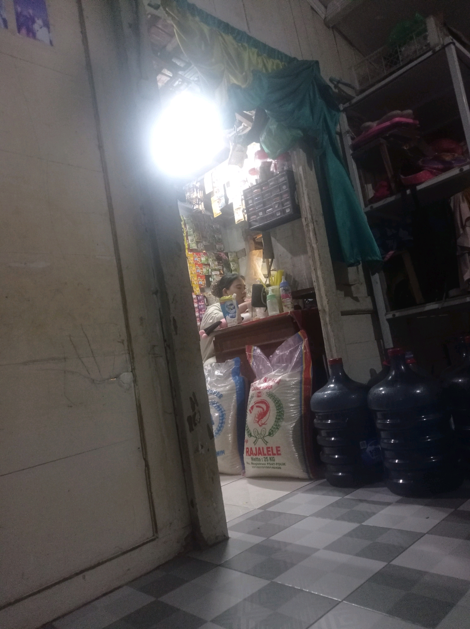 This is a low-light photograph of the interior of a small shop, possibly a convenience store or warung, located in Babalan, Indonesia. The scene is dimly lit, with a bright light bulb hanging from the ceiling creating strong shadows and glare.
In the background, a person is partially visible behind a counter, wearing a light-colored t-shirt. They appear to be interacting with something on the counter, possibly a product or customer. Several shelves are visible behind the counter, stocked with various items, including what appear to be packaged goods and possibly cleaning supplies.
In the foreground and to the right of the frame, several large, dark blue water jugs are lined up on the floor. To the left of the water jugs and in front of the counter, there are two large white plastic bags, one of which has a red and green logo that reads "RAJALELE" and "25 KG". The floor is tiled with a black and white checkered pattern. The overall environment suggests a small, potentially cluttered retail space.