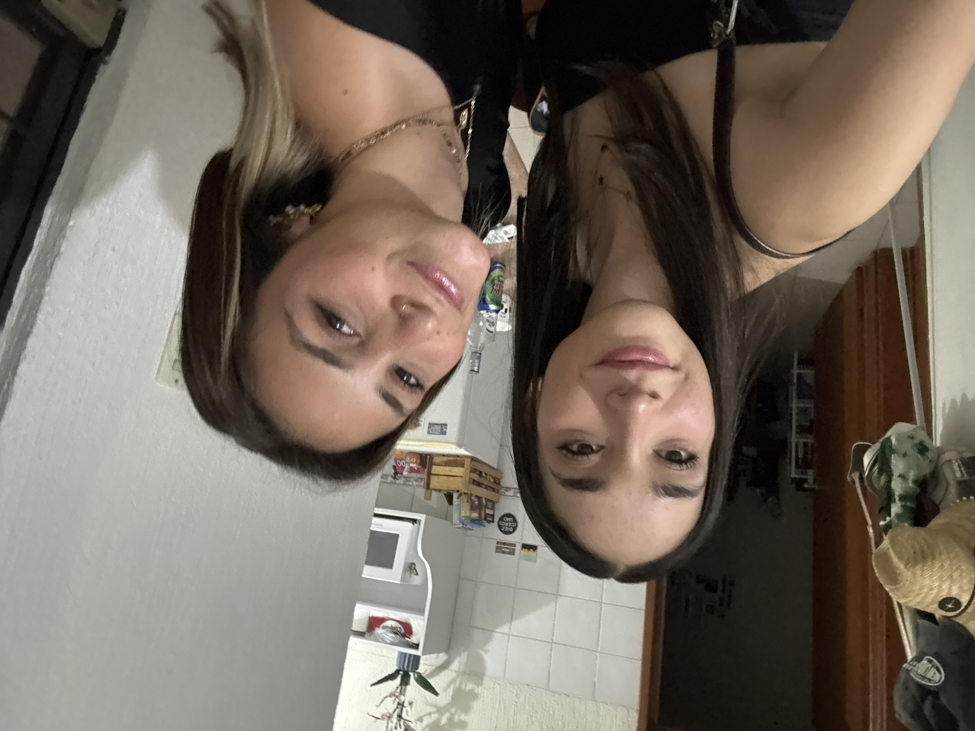 Two women are posing for a selfie indoors. The camera is angled downward. The woman on the left has light brown hair with blonde highlights. She is wearing a black top and a gold necklace, and she has a nose ring. The woman on the right has long, dark brown hair. She is wearing a black top with spaghetti straps and a silver necklace.
In the background, there is a microwave, shelving, a tiled wall, and a doorway. A tripod is in the lower center of the frame. A doorway, hats and coats on a coat rack, and a doorframe are visible on the right side of the frame. Based on the appearance of the scene and the presence of a brand of beer sold locally, the location is in San Andrés Cholula, Mexico.