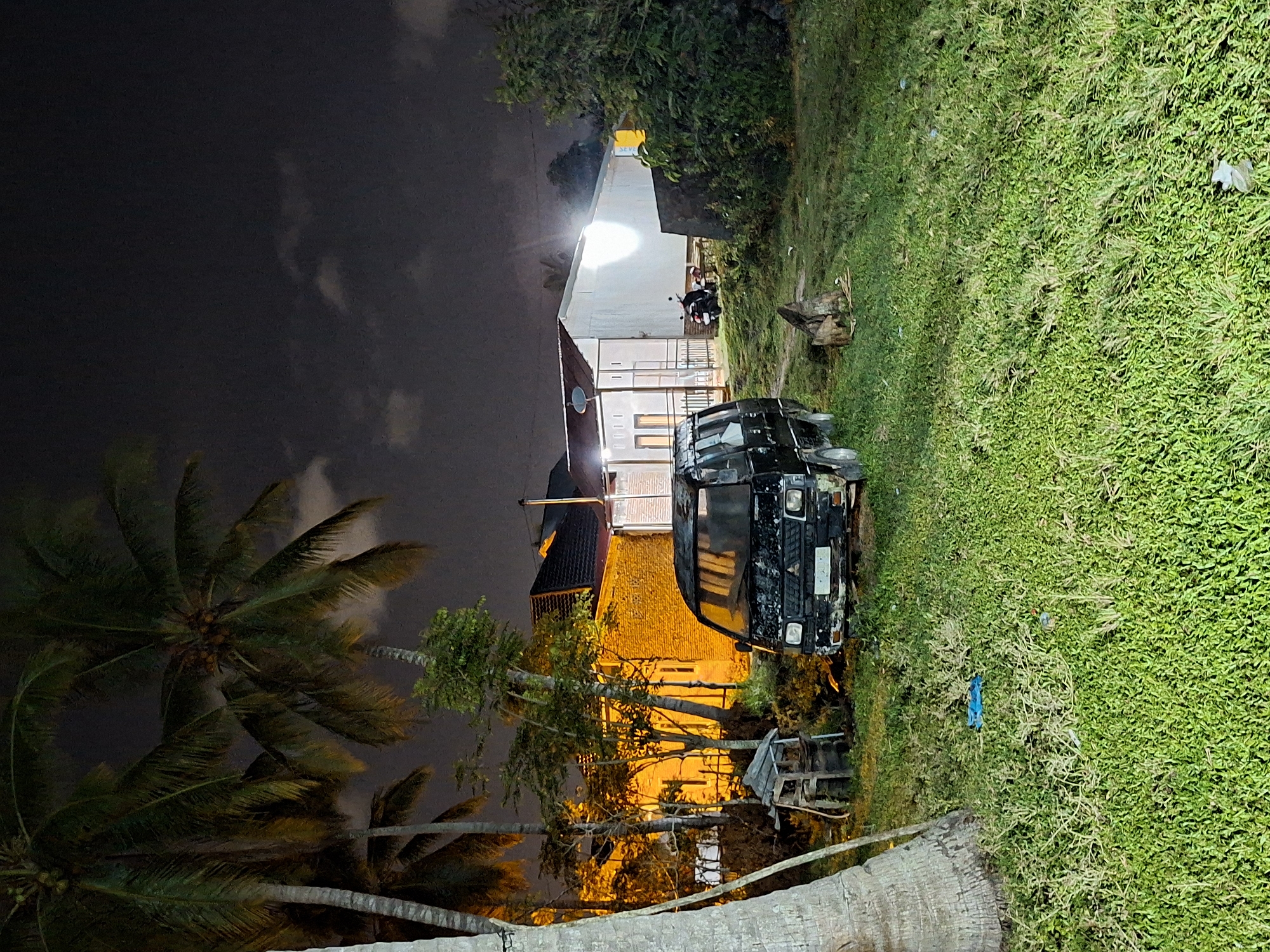 At night in Lhokseumawe, Indonesia, a black truck is parked on a grassy area adjacent to a brightly lit house. Palm trees are visible against the dark sky. A motorcycle is parked near the house. The environment suggests a residential area.