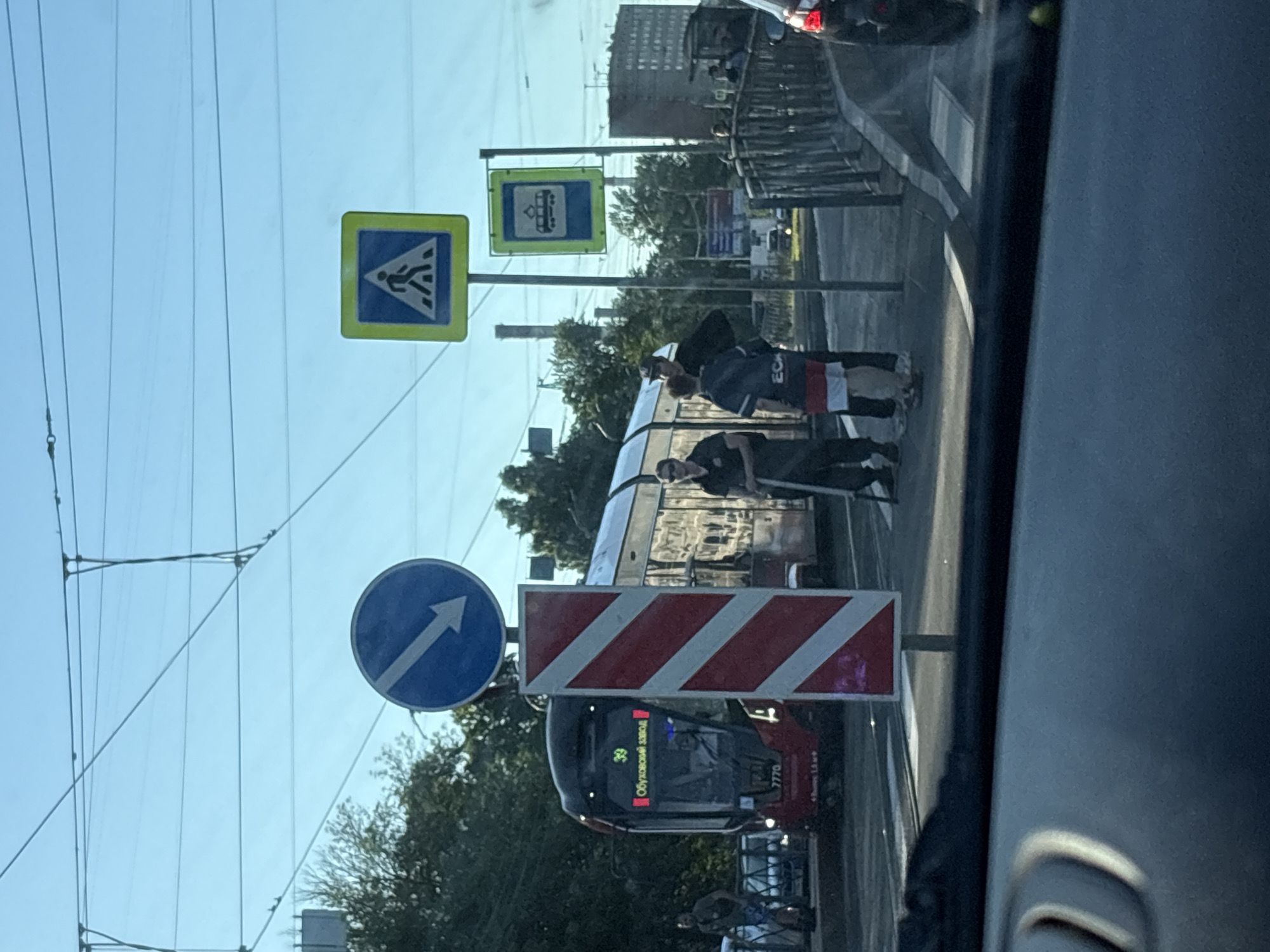 This image depicts an urban scene in Saint Petersburg, Russia, likely along the route of **Tram 39 near Obukhovsky Zavod**, an industrial area in the city's south-east. The perspective is from inside a vehicle, looking forward through the windshield on a clear, sunny day.
In the mid-ground, a modern red and black low-floor tram, identified as **Tram 39** with the destination "Обуховский завод" (Obukhovsky Zavod) and number "7770," is prominently featured on tracks to the left. The tram appears to be stopped or moving slowly. A large red and white diagonal striped barrier is positioned centrally in the foreground, partially obscuring the tram. Above this barrier is a blue circular road sign with a white arrow pointing diagonally downwards to the right.
To the right of the tram, a group of people is visible. Two men are standing near the tram tracks; one wears sunglasses and a dark shirt, holding a white stick or pole, while the other, wearing a dark shirt with "ECL" visible and striped shorts, stands beside him. Further right, a metal-railed bridge or overpass stretches into the background, with several indistinct figures on it.
Additional road signs are present: a square yellow sign with a black pedestrian symbol (indicating a pedestrian crossing) is high on the right, and below it, a blue square sign with a white tram icon (indicating a tram stop). The background features green trees, residential or industrial buildings, and overhead power lines against a bright blue sky. A partial view of another vehicle is visible on the far right. The dark dashboard of the observer's vehicle is visible in the immediate foreground.