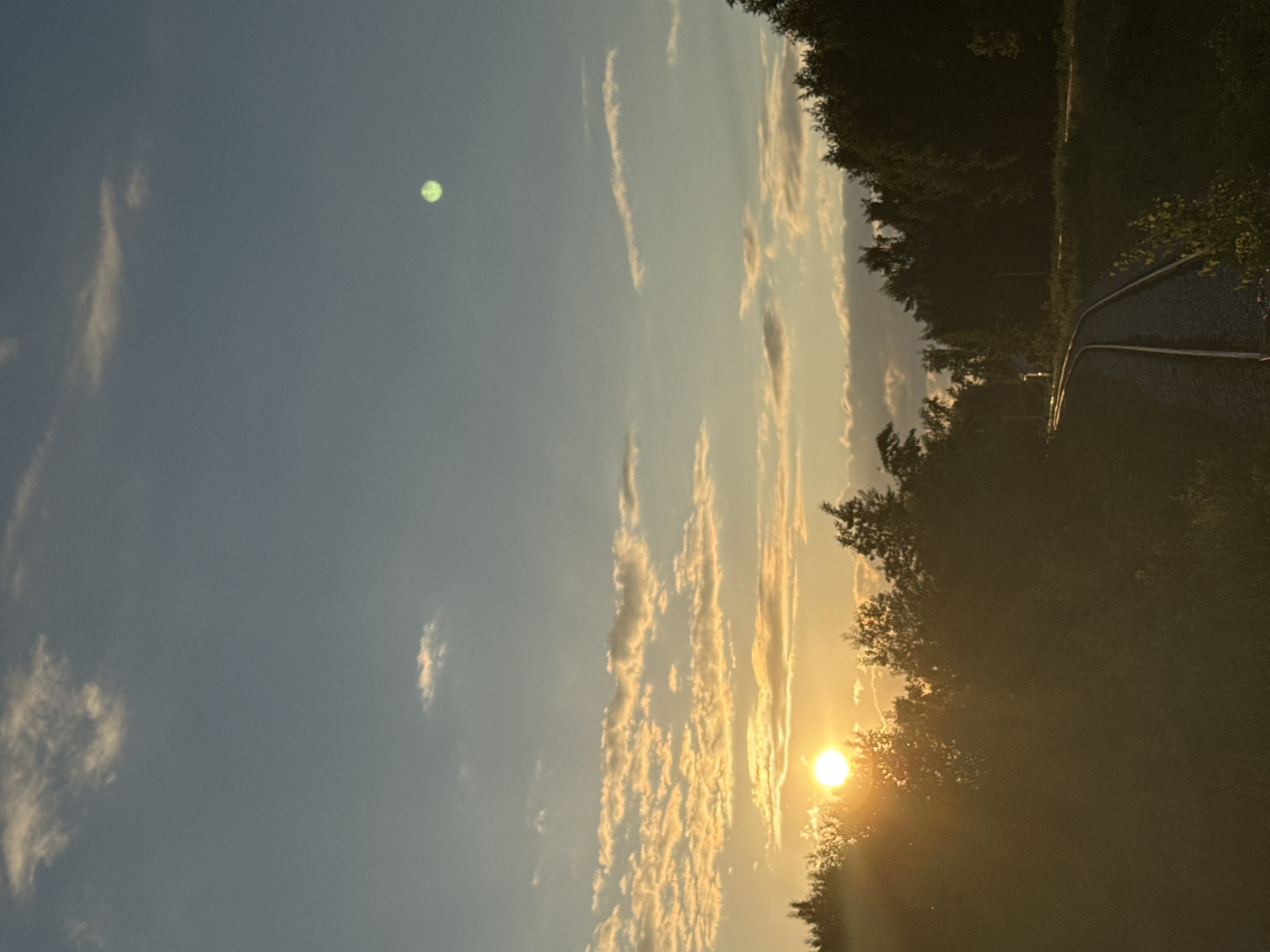 The image depicts a sunset over a railway track in Chaikovsky, Russia. The sun, a bright orb, is partially obscured by trees and clouds, casting a warm, golden glow across the scene. The sky is a gradient of blue and orange, with scattered clouds catching the last rays of sunlight. The railway track curves into the distance, bordered by dense foliage and trees on either side. A faint green light is visible in the upper right portion of the sky. The overall impression is one of a serene, natural landscape at dusk.