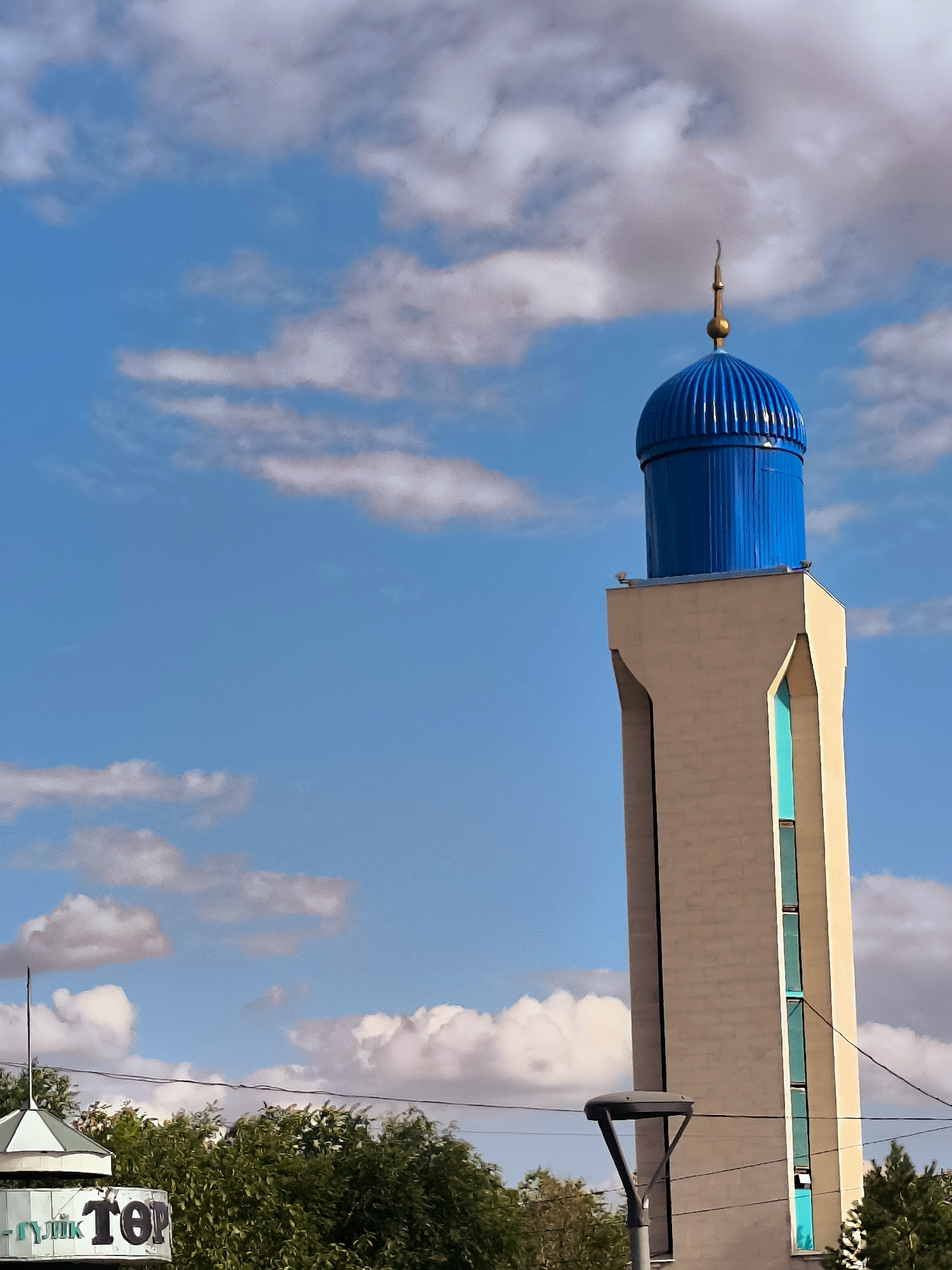 The image shows a close-up view of a minaret against a blue sky with scattered clouds. The minaret is tall and slender, constructed from light-colored material. It features a striking blue dome at the top, topped with a golden spire. There are narrow, vertically oriented turquoise windows along one side of the minaret. In the foreground, partially visible at the bottom left, is a portion of a building with signage that appears to be in Cyrillic script, and some green trees. A street lamp is also visible in the lower right quadrant of the image. The scene is set outdoors, under daylight conditions. The minaret is located in the city of Qarağandı, Kazakhstan, in front of the city administration building.