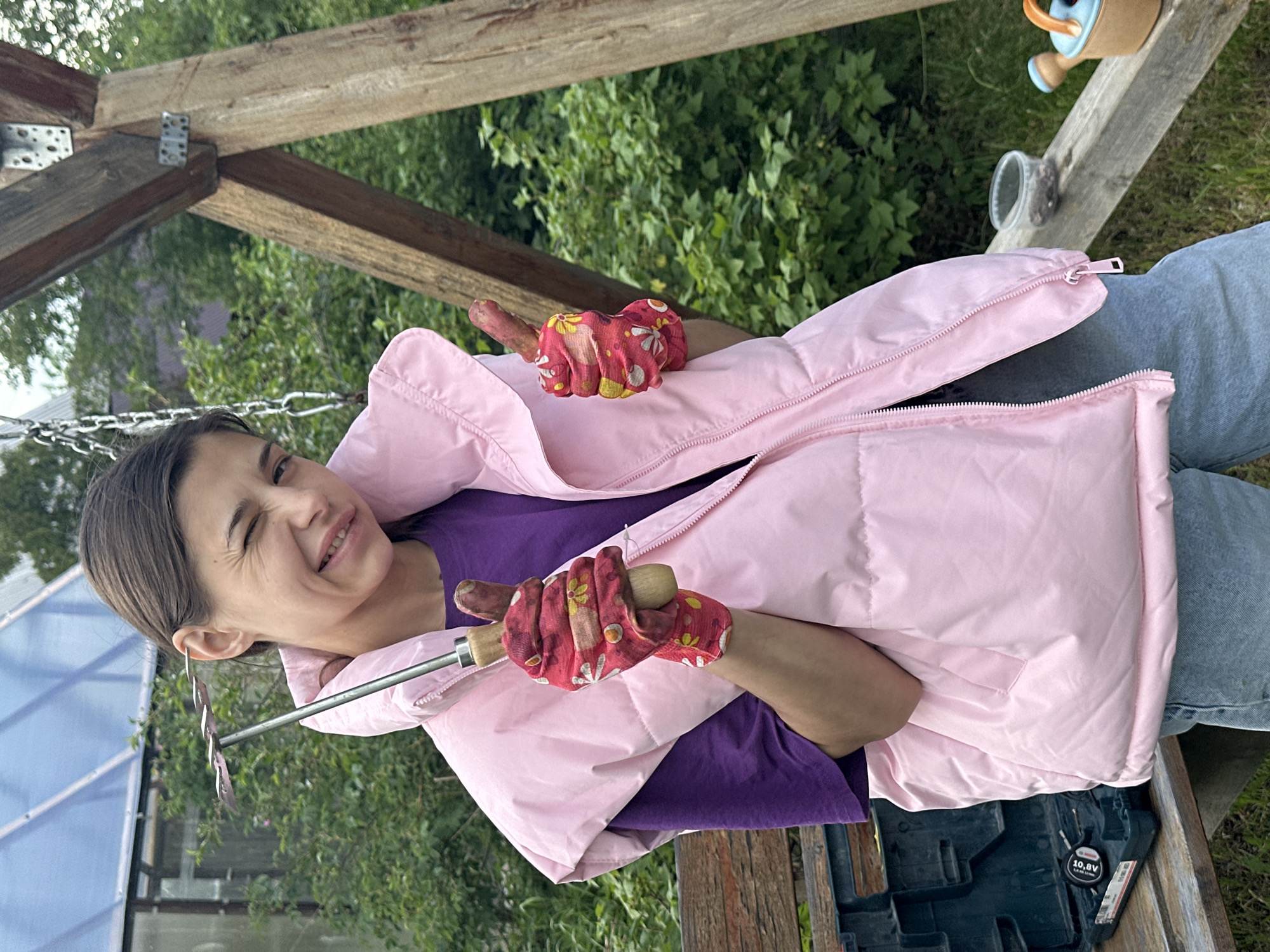 A woman in Khokhryaki, Russia, holds gardening tools. She wears pink gloves and a pink, unzipped puffer jacket over a purple shirt and jeans. The background includes a wooden structure, possibly a greenhouse or shed frame, with greenery visible beyond. A watering can and other items sit on a nearby wooden surface.