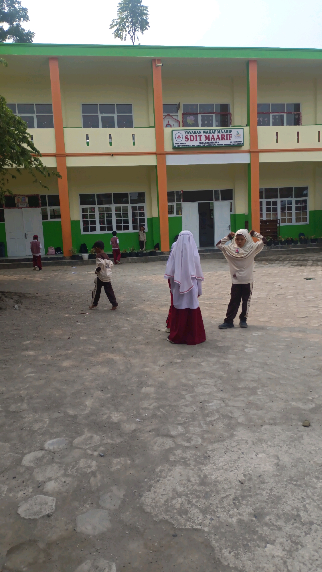 The image shows the exterior of a school building, identified by a sign as "SDIT MAARIF" located in Padang Panjang, Indonesia. The school building has a yellow facade with green and orange trim. There are several windows on both levels of the building, and the entrance to the school is visible in the center.
In the foreground, on a paved courtyard, several children are present. Three children are the most prominent: one in the center left is wearing a light-colored shirt and dark pants, walking with their head down. To the right of center, a child wearing a white hijab and a dark red skirt is standing, facing away from the camera. To the right of this child, another child, also wearing a light-colored hijab and shirt with dark pants, is striking a pose, with their hands on their head and their arms bent.
Other children are visible in the background, closer to the school building, including some standing near the entrance and one near the left side of the building. The courtyard appears to be made of concrete and gravel, with some scattered debris. The sky is overcast, and trees are visible behind the school building. The overall scene depicts a school environment with children engaged in outdoor activities.