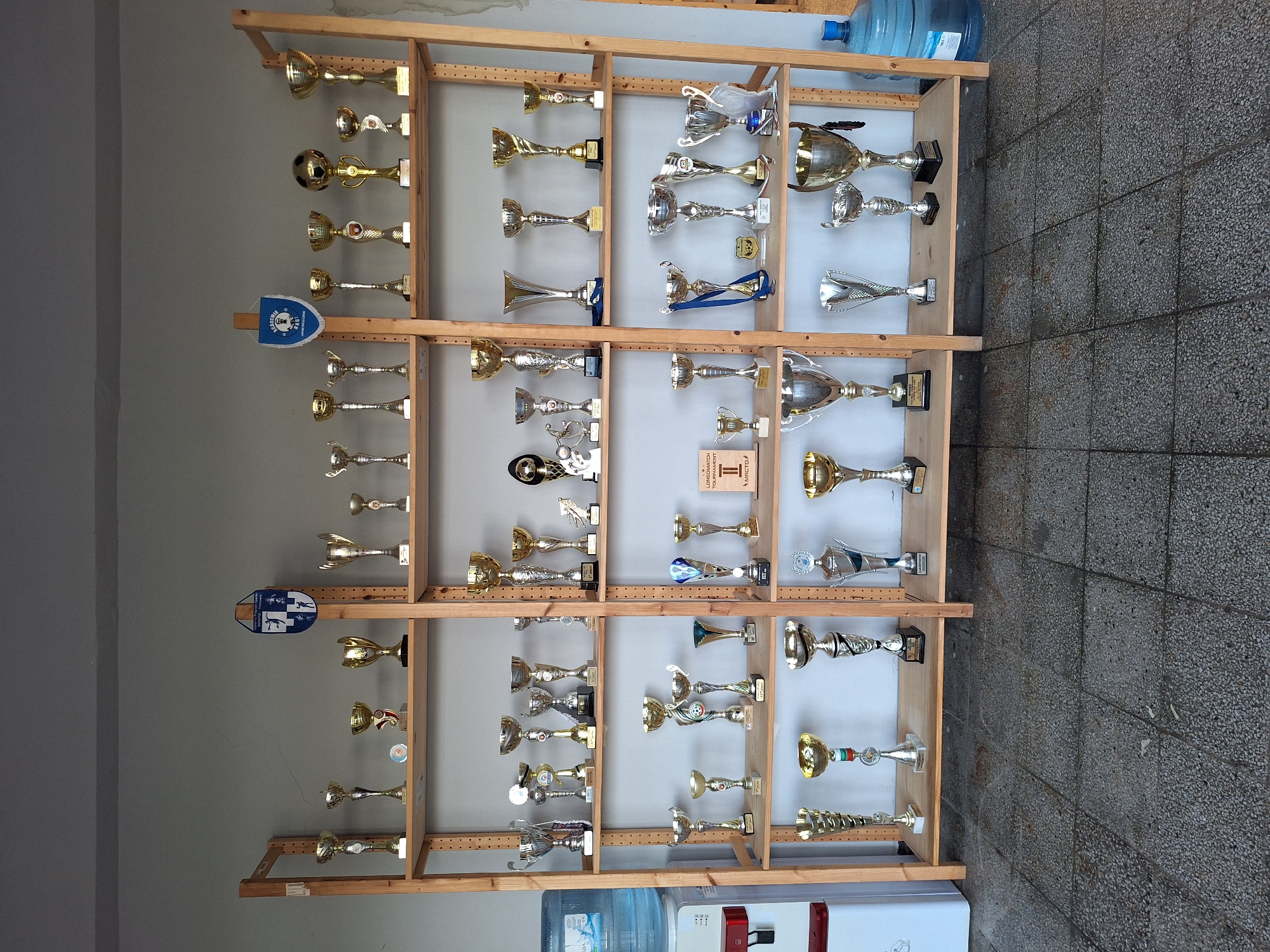 The image depicts an indoor scene in Ezerovo, Bulgaria, showcasing a multi-tiered wooden shelving unit displaying approximately 60-70 sports trophies. The unit, constructed from light-colored wood, comprises four vertical support sections and five horizontal shelves positioned against a plain, light-grey wall.
The trophies are diverse in design, predominantly metallic with gold, silver, and bronze-toned finishes. They include classic cup shapes, figurines (such as a stylized footballer), and spherical tops resembling a football. Many trophies rest on dark bases, some with legible inscriptions.
Specific elements on the shelves include:
* On the upper left shelf, a blue and white shield-shaped pennant is partially visible, bearing the Cyrillic text "СПОРТНА АКАДЕМИЯ БОРИСЛАВ" (Sports Academy Borislav).
* On the upper right shelf, a blue and white triangular pennant with a crest is displayed, reading "АКАДЕМИЯ НАЦИ" (Academy Naci).
* On a lower shelf, a small brown rectangular plaque with "LONGOMATCH" and "II МЯСТО" (2nd Place) is present.
* One trophy on a middle shelf features a blue ribbon attached.
To the far left, a white water dispenser with a red spigot is visible, topped by a large, clear blue 5-gallon water bottle labeled "AQUA." To the far right, a similar blue 5-gallon water bottle rests on the floor.
The floor is covered with dark grey, speckled concrete or terrazzo tiles, showing visible grout lines and signs of wear. The lighting is consistent with indoor ambient conditions. No individuals are present in the scene.