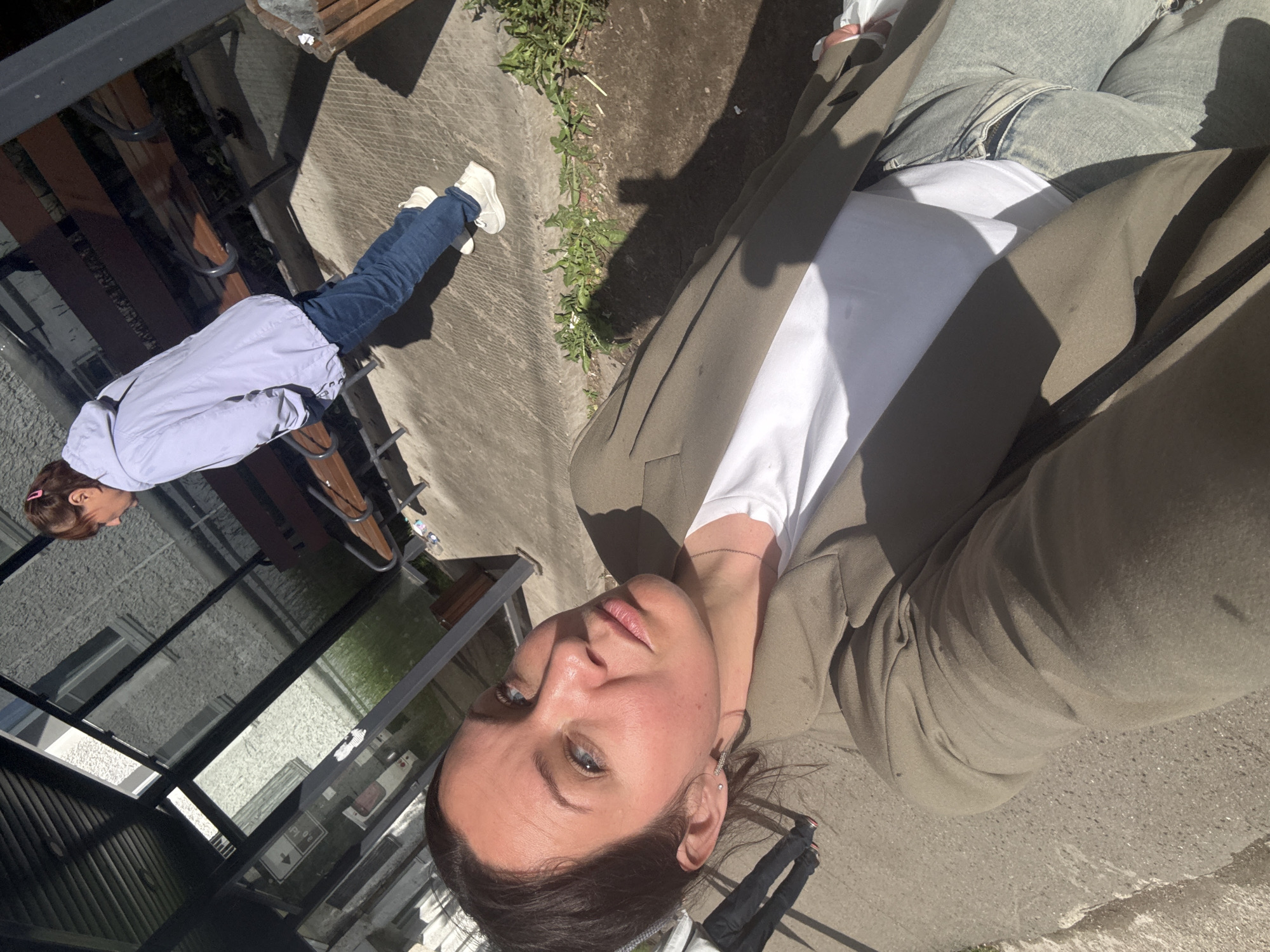 The image captures an outdoor scene, likely in an urban or suburban setting, under bright sunlight. The primary subject is a woman taking a selfie, visible in the foreground, angled upwards. She has dark hair, blue eyes, and a neutral to slightly contemplative expression. She is wearing a white t-shirt layered under a khaki or olive-green blazer, with light-wash jeans partially visible. Strong sunlight casts distinct shadows on her face and clothes.
In the mid-ground, a second person is partially visible to the left (when the image is viewed correctly oriented). This individual is seen from the chest down, wearing a light blue or lavender long-sleeved top or jacket, dark blue jeans, and white sneakers. They appear to be standing or seated near a dark-framed wooden bench, facing away from the camera.
The background consists of a paved surface, likely concrete or asphalt, with patches of small green plants or weeds growing in cracks. Reflections of a building facade and sky are visible in a dark, possibly glass or polished, surface to the left. The scene suggests a casual outdoor environment, possibly a walkway, patio, or park area. No discernable text is visible within the image. The time of day appears to be sunny, creating sharp contrasts and highlights.