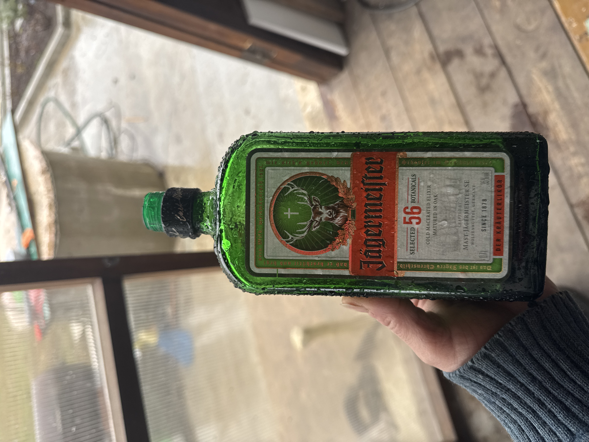 A hand holds a bottle of Jägermeister. The bottle is green, wet with condensation, and features the brand's iconic logo with a deer head and a cross. The label includes text such as "56 Botanicals," "Cold Macerated Elixir," and "Der Kräuterlikör." The person holding the bottle wears a dark blue knitted sweater. The background shows a blurry outdoor scene with wooden frames and a hint of foliage. There's no indication that this image was taken in the city of Chaikovsky, Russia.