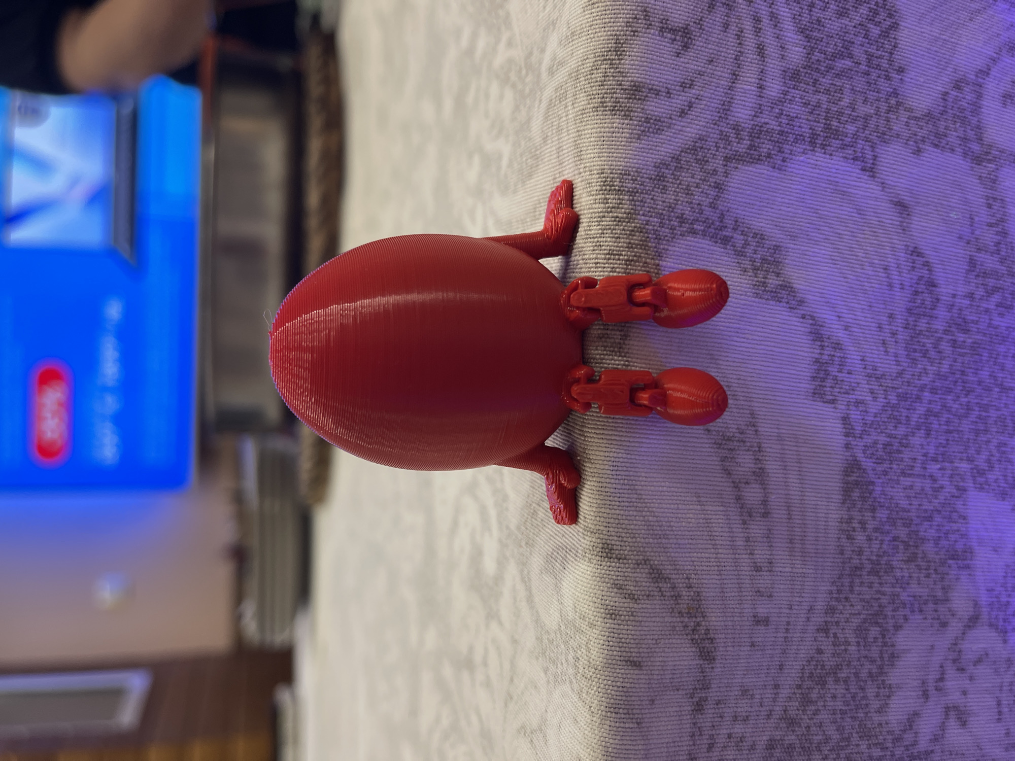 A red, egg-shaped object with articulated limbs is resting on a patterned surface that appears to be a tablecloth or rug. The background shows a television displaying a blue screen with white text and a red logo, along with a blurred glimpse of a room. The location is not discernible from the given image.