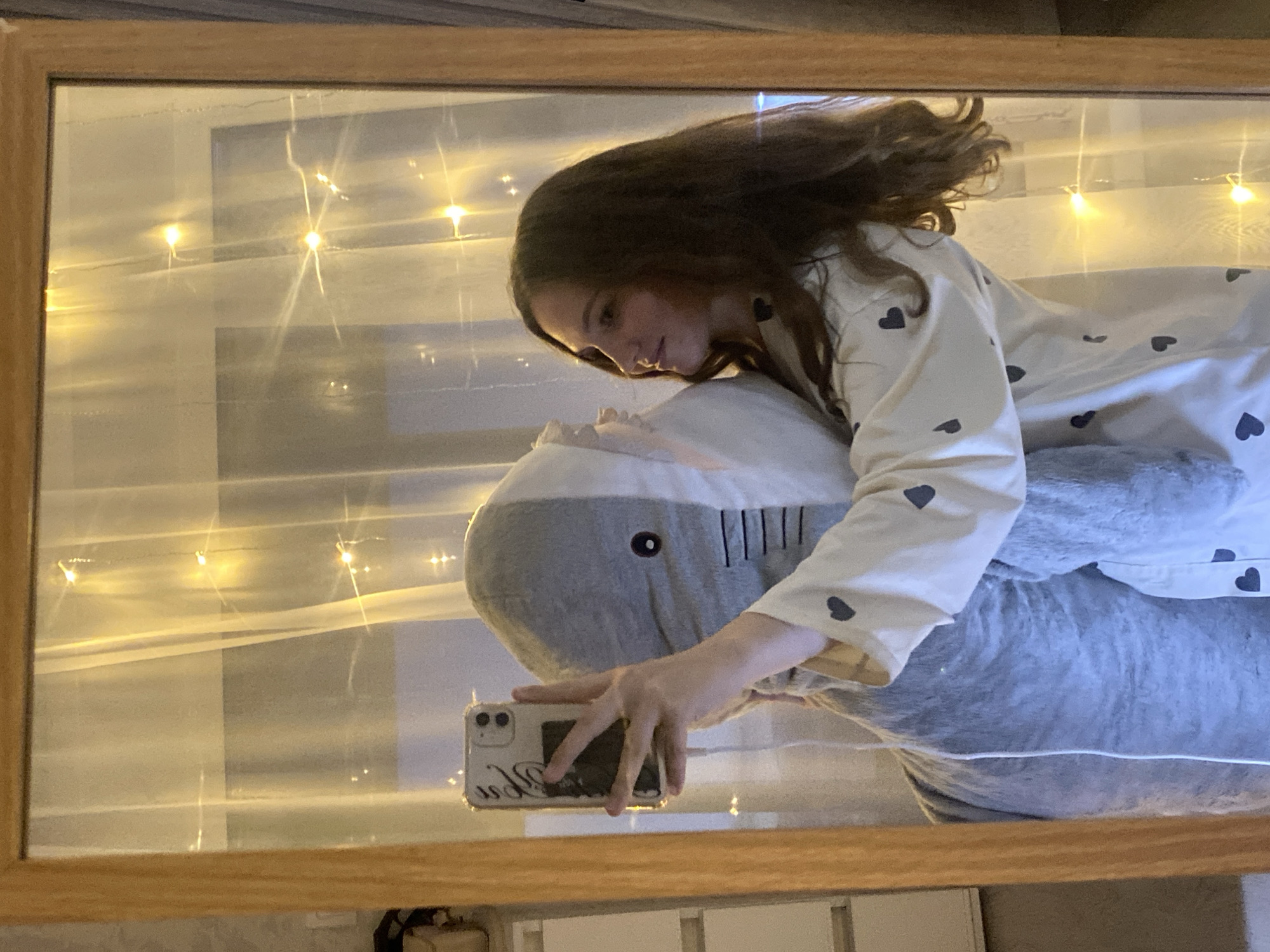 A mirror reflects a person with long brown hair wearing a light-colored pajama top with dark hearts lying on a large grey plush shark. The person is holding a mobile phone to take a photo. Behind the person and shark are strands of decorative string lights. The mirror has a light-colored wooden frame. The reflection shows a window behind the person. It is not possible to determine if this scene is in Izhevsk, Russia.