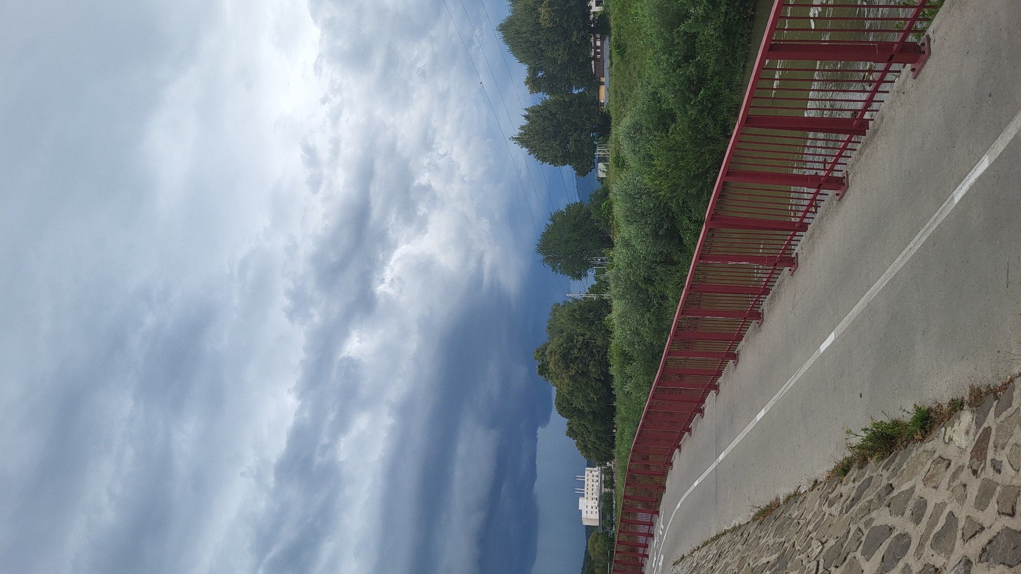 The image depicts a cloudy sky over a waterfront path and a red railing in the city of Prešov, Slovakia. The path, made of asphalt, is marked with a white line and runs alongside a stone embankment on the left. The red railing separates the path from a body of water on the right. Beyond the water, lush green trees and bushes line the riverbank. In the background, a large white building with multiple windows and smokestacks is visible, suggesting an industrial or institutional complex. The overall scene conveys an atmosphere of an impending storm, with dark, heavy clouds dominating the sky.