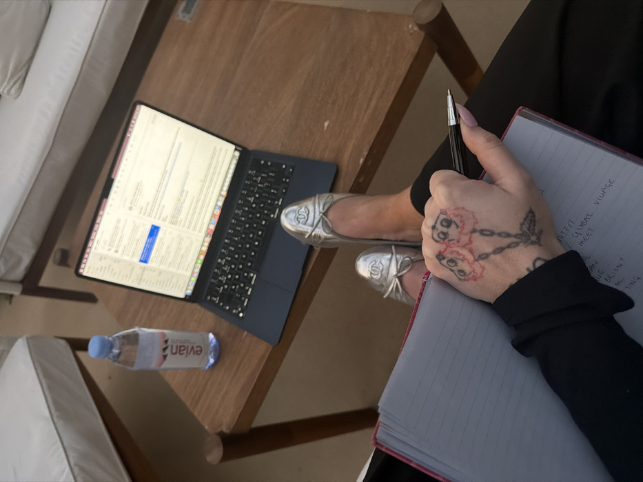 An overhead photograph depicts a person's lower body and left arm engaged in an activity within an outdoor or semi-outdoor setting in Dubai, United Arab Emirates. The individual wears black trousers and silver Chanel ballet flats. Their left hand, tattooed with two stylized, smiling red flowers connected by a chain-like stem, holds a black pen over an open notebook with light blue lined pages and a red spine. Partial handwritten text visible includes "OUTFIT," "GLOBAL VILLAGE," and "MEET." On a wooden table directly in front, a dark-colored laptop is open and displaying an email application interface. To the laptop's left, a clear plastic Evian water bottle with a blue cap is positioned. In the blurred background, elements of white-cushioned wooden lounge furniture are visible, suggesting a patio or terrace environment.